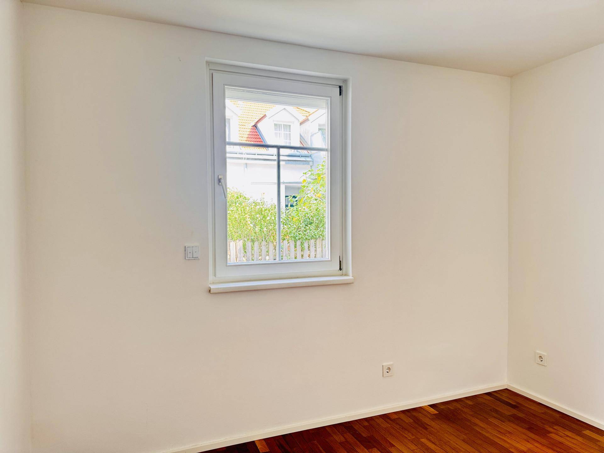 Helles Zimmer mit Parkettboden und Fenster, das einen Blick auf die Nachbarschaft ermöglicht.