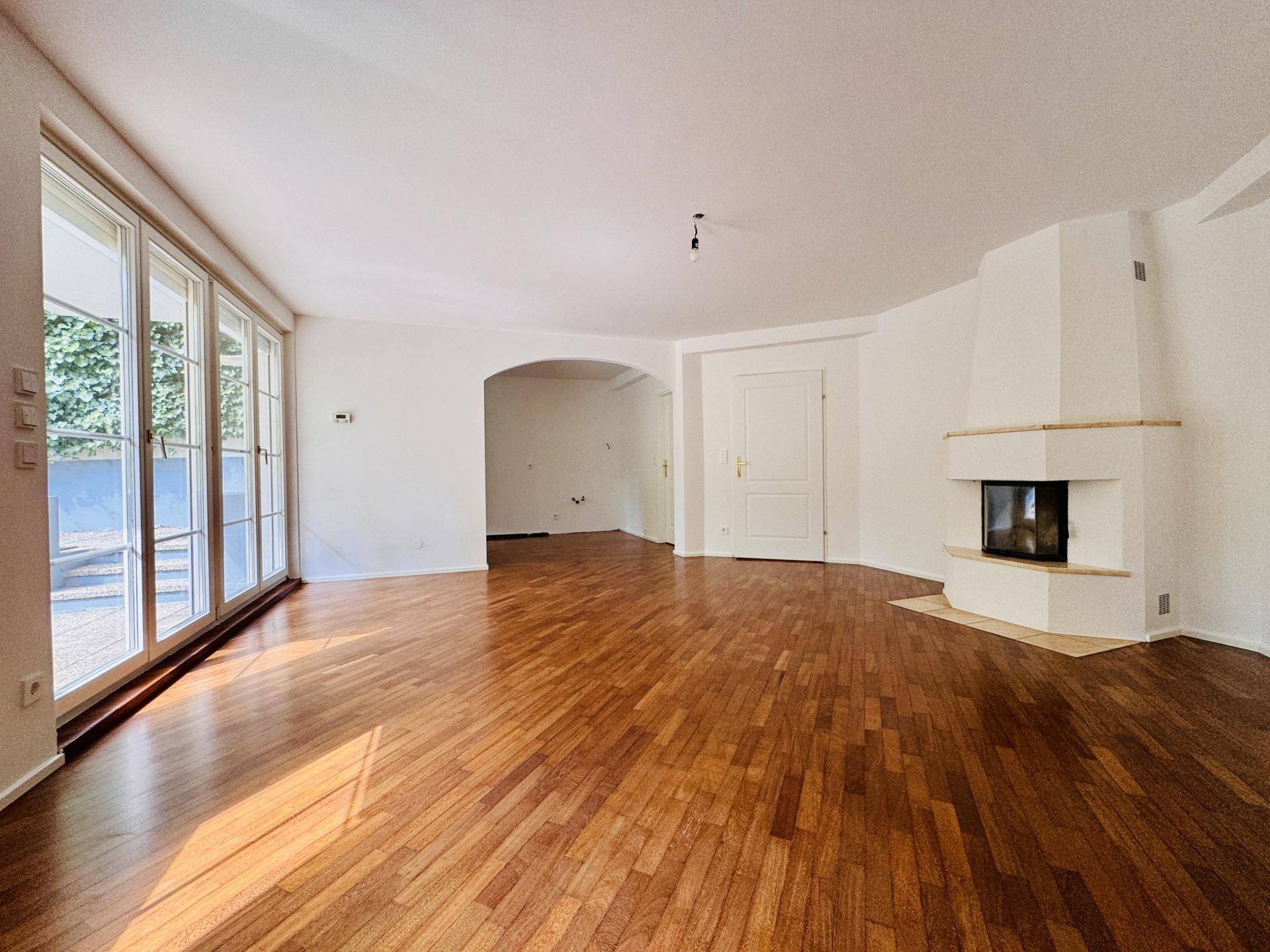 Helles Wohnzimmer mit Parkettboden, Kamin und Zugang zu weiteren Räumen durch einen Bogen.