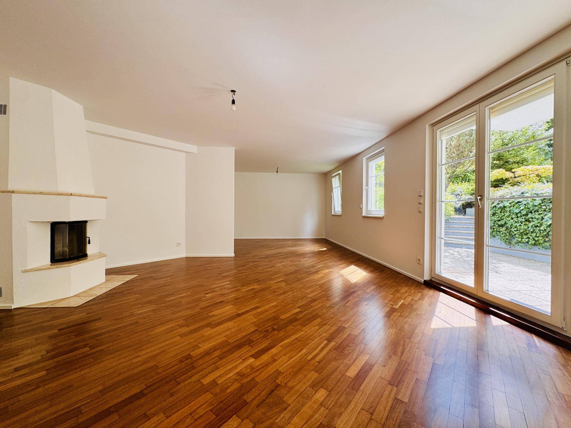 Geräumiges Wohnzimmer mit hellem Parkettboden, Kamin und großen Fenstern zum Garten.