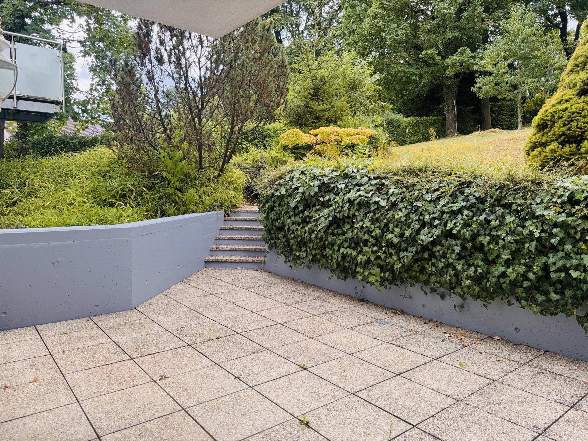 Geräumige Steinterrasse mit Treppenaufgang zum Garten, umgeben von üppigem Grün.