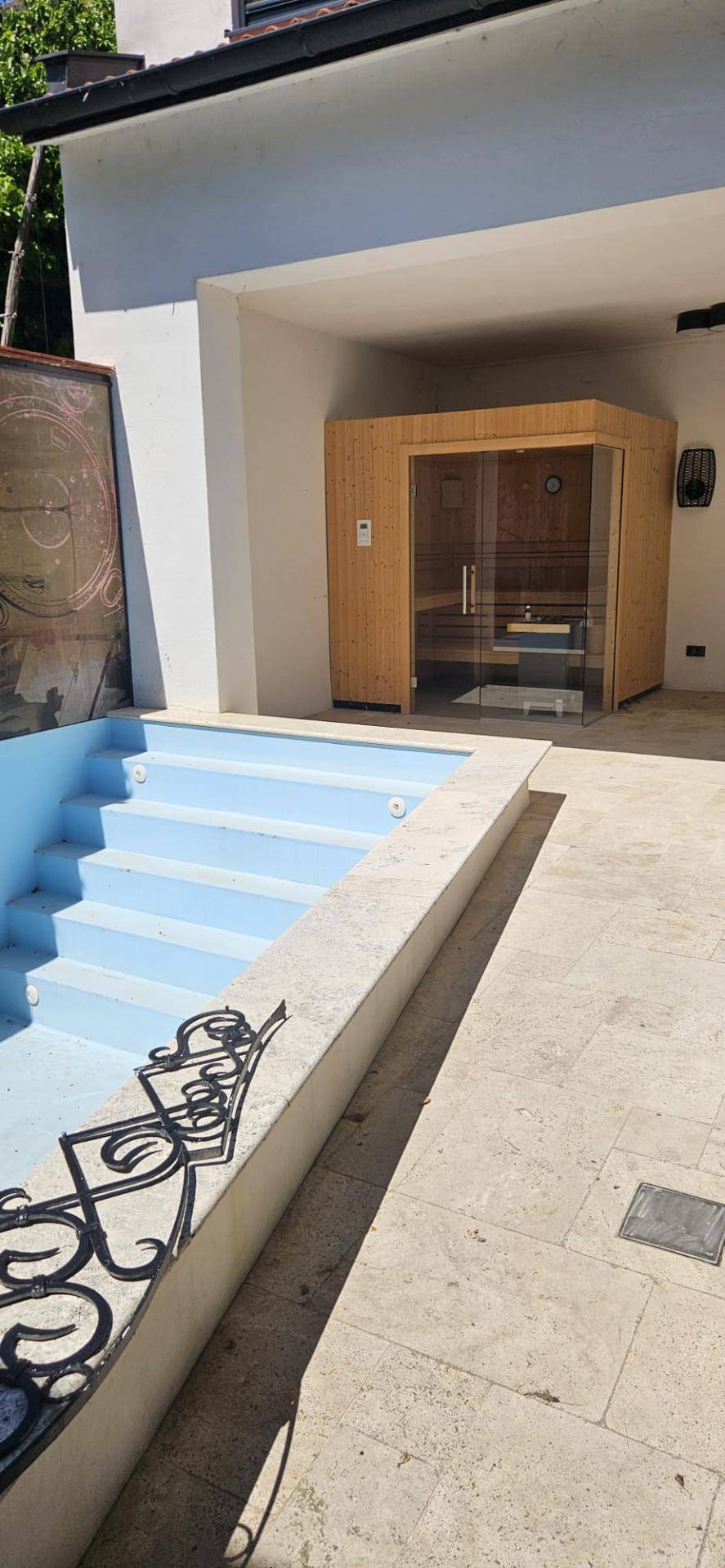 Außenbereich mit einem blauen Pool und einer Holzsauna, umgeben von hellen Steinplatten.