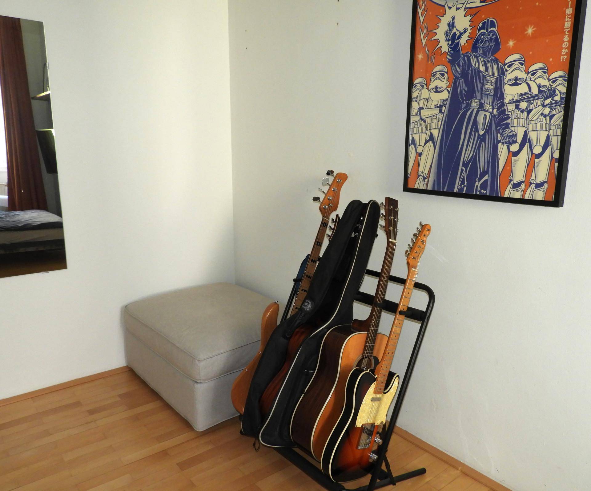 Ecke eines Zimmers mit mehreren Gitarren auf einem Ständer und einem Poster an der Wand.