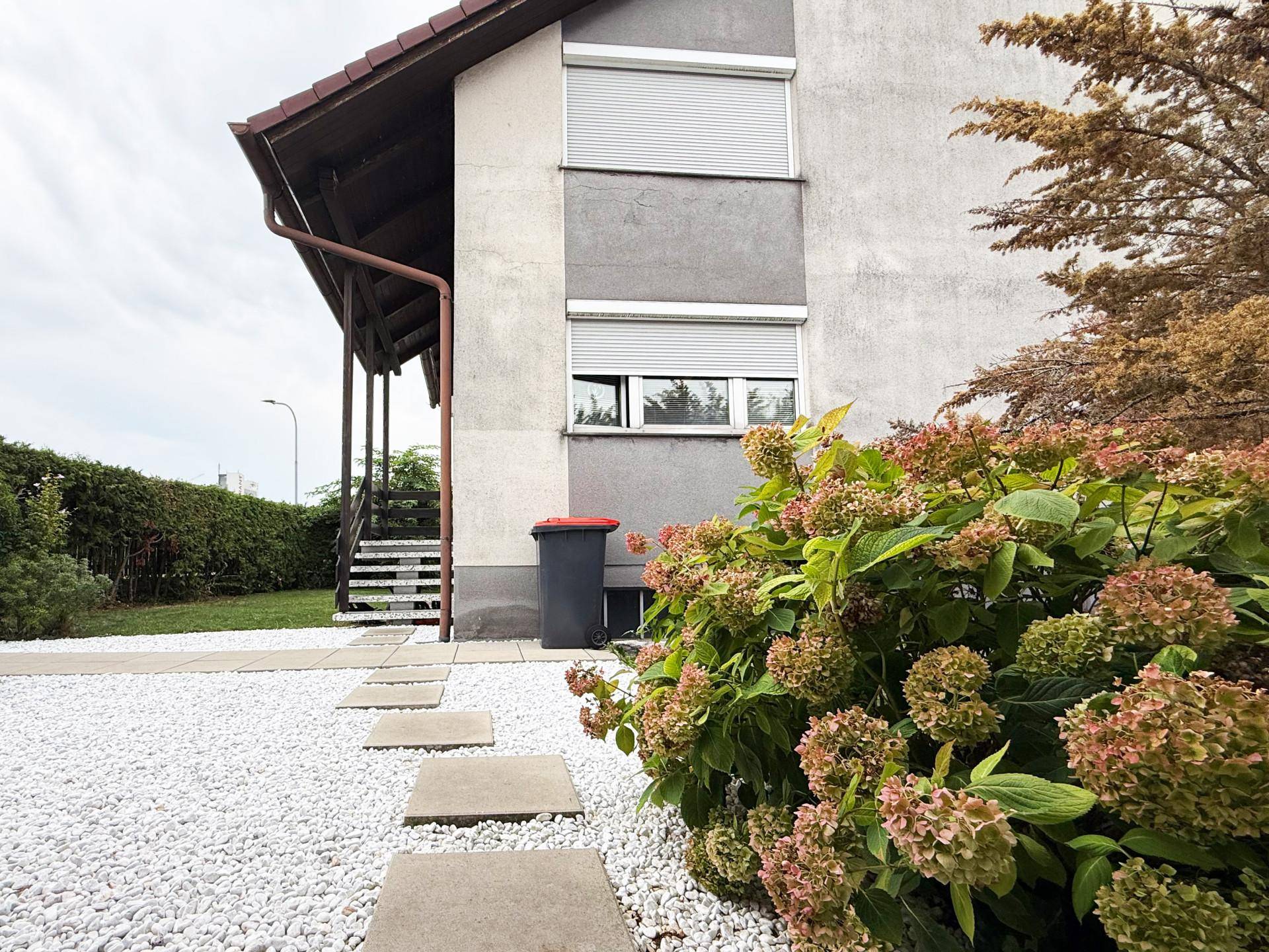 Seitenansicht des Hauses mit Kiesweg, Blumenbeet und Rollläden an den Fenstern.
