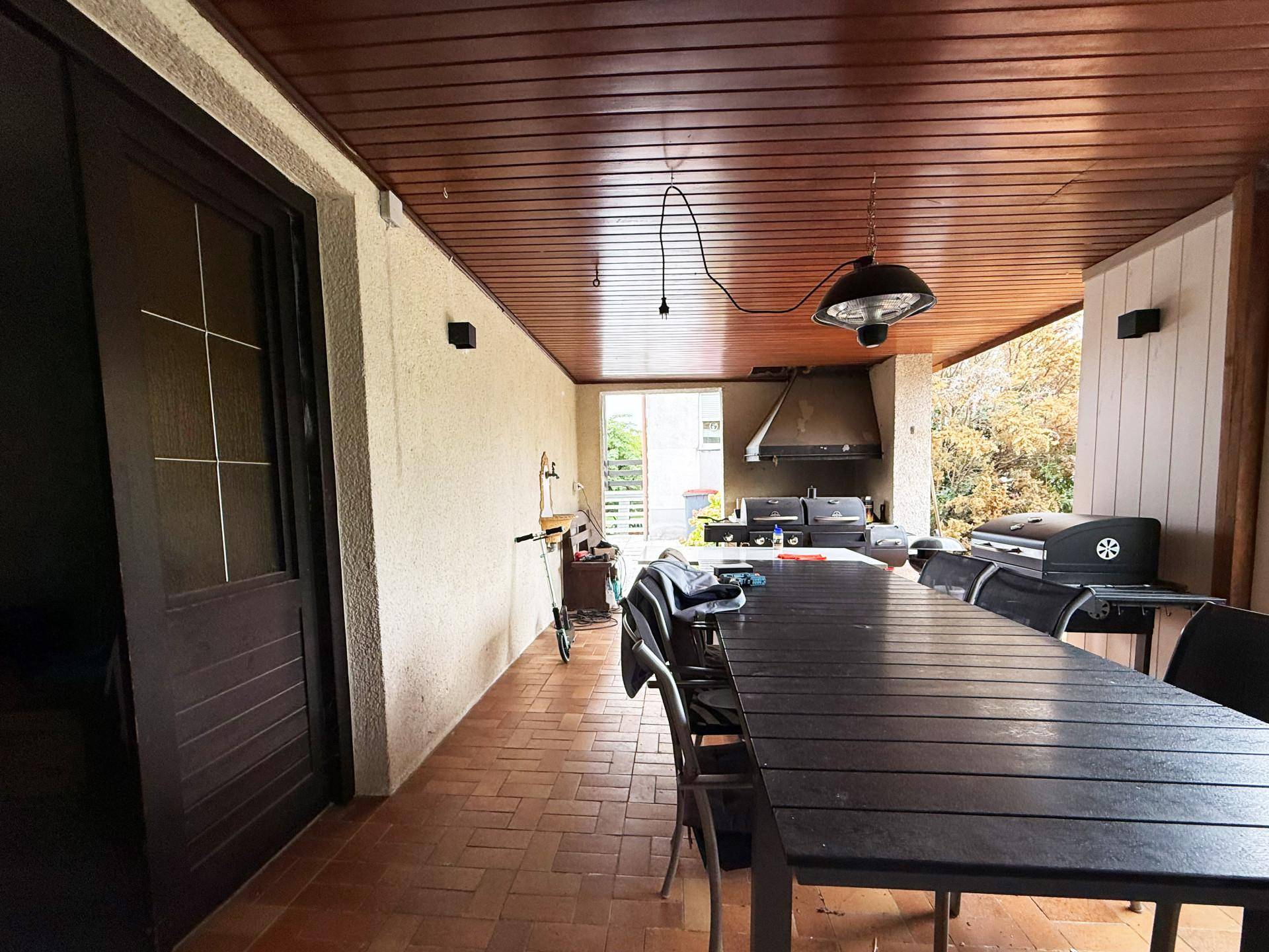 Überdachte Terrasse mit großem Esstisch, Grillbereich und Holzdecke.