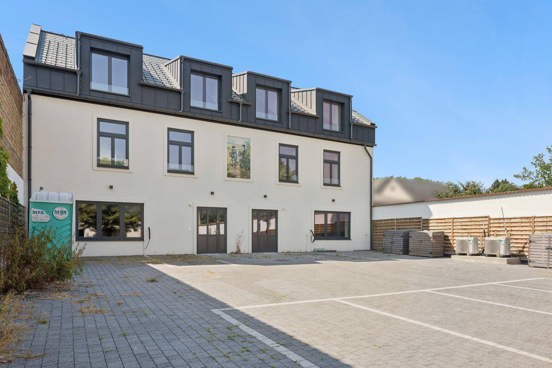 Seitenansicht des Gebäudes mit Parkflächen, einem provisorischen Zaun und moderner Architektur.