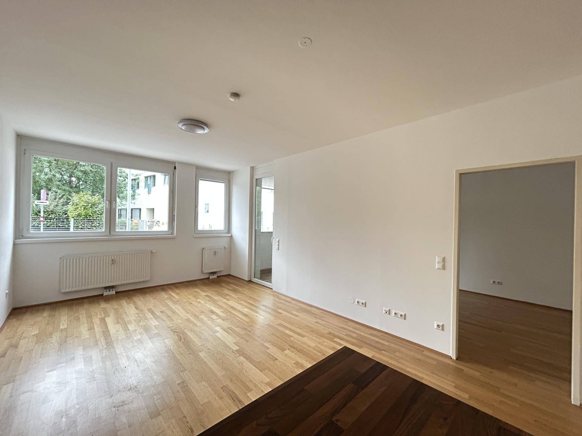 Geräumiges Wohnzimmer mit hellem Holzboden und großen Fenstern, die viel Tageslicht hereinlassen.
