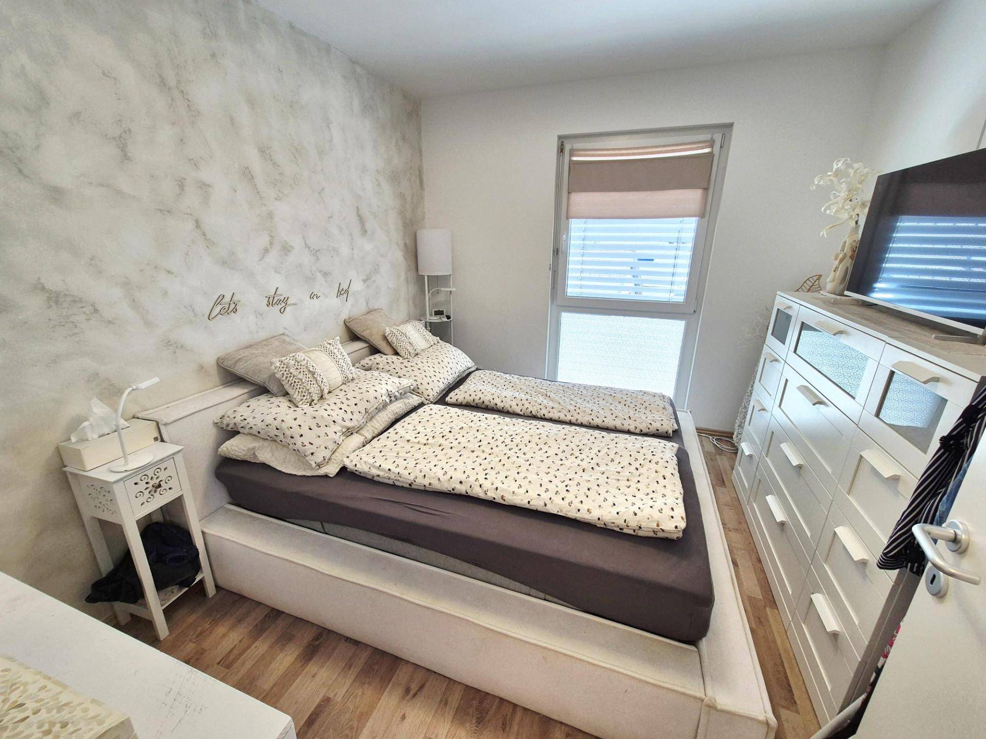 Helles Schlafzimmer mit Doppelbett, Kommode und Fenster für natürliches Licht.