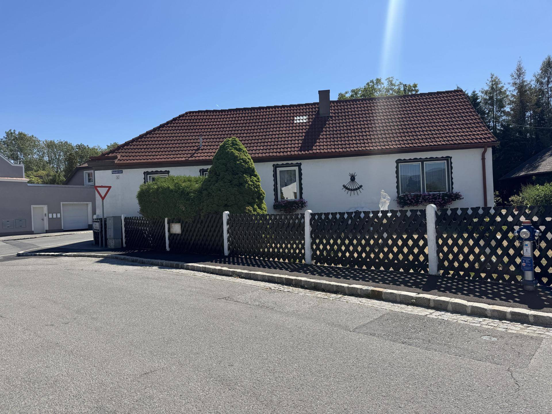 Ansicht eines weißen Hauses mit rotem Ziegeldach, dunklem Zaun und einer ruhigen Straße im Vordergrund.