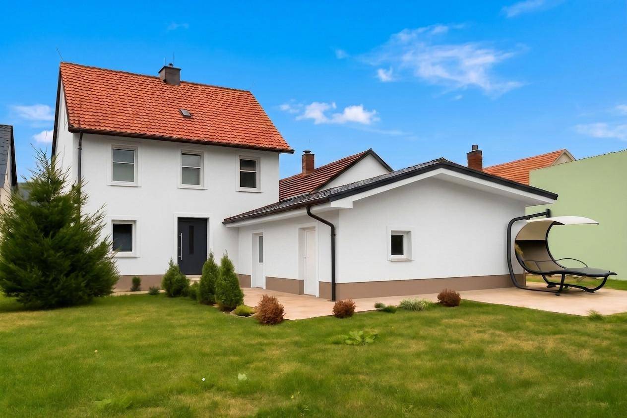 Weißes Einfamilienhaus mit rotem Ziegeldach, großem Rasen und einer modernen Liege im Gartenbereich.