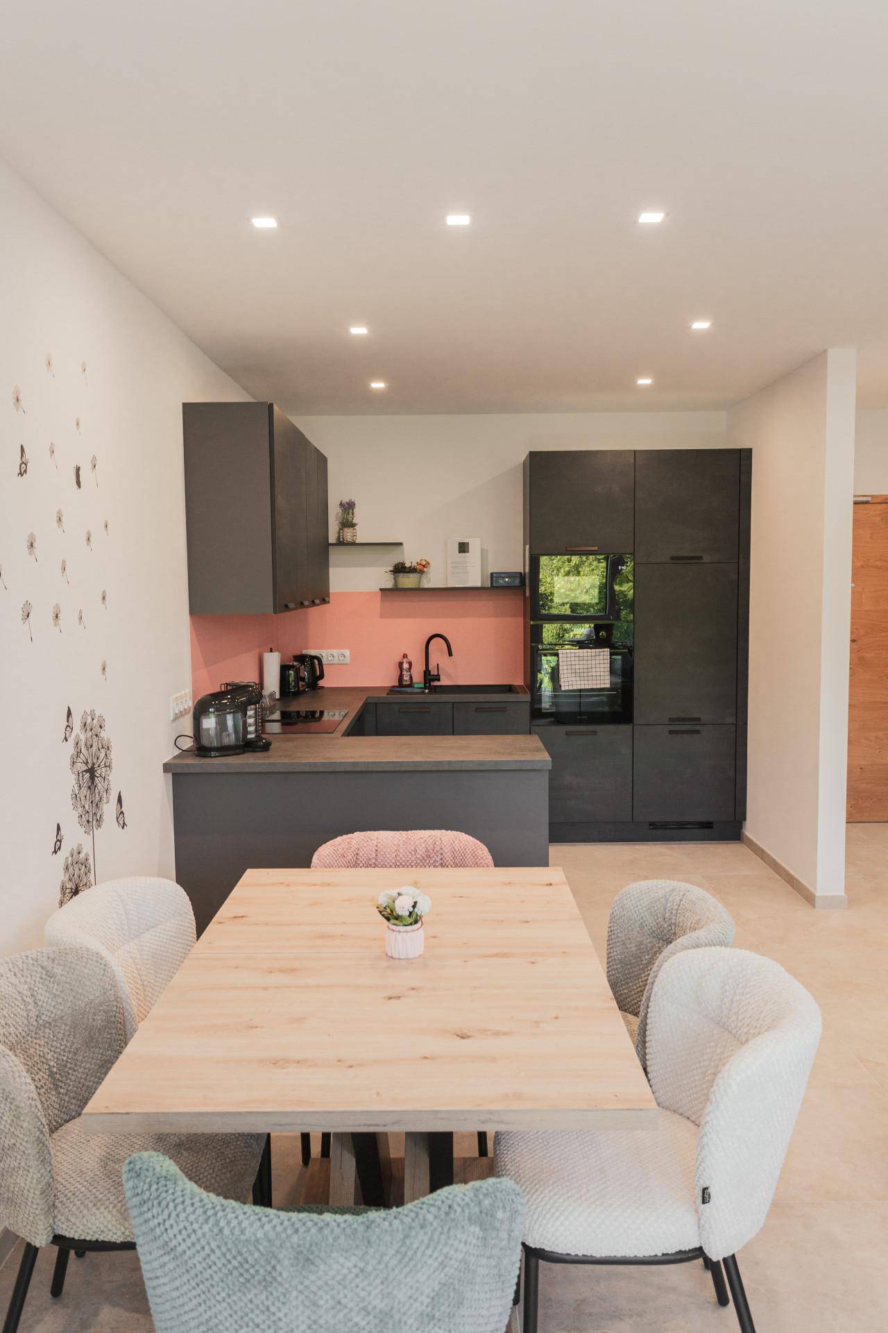 Moderne Küche mit dunklen Schränken, rosa Rückwand und hellem Holztisch für gemeinsame Mahlzeiten.