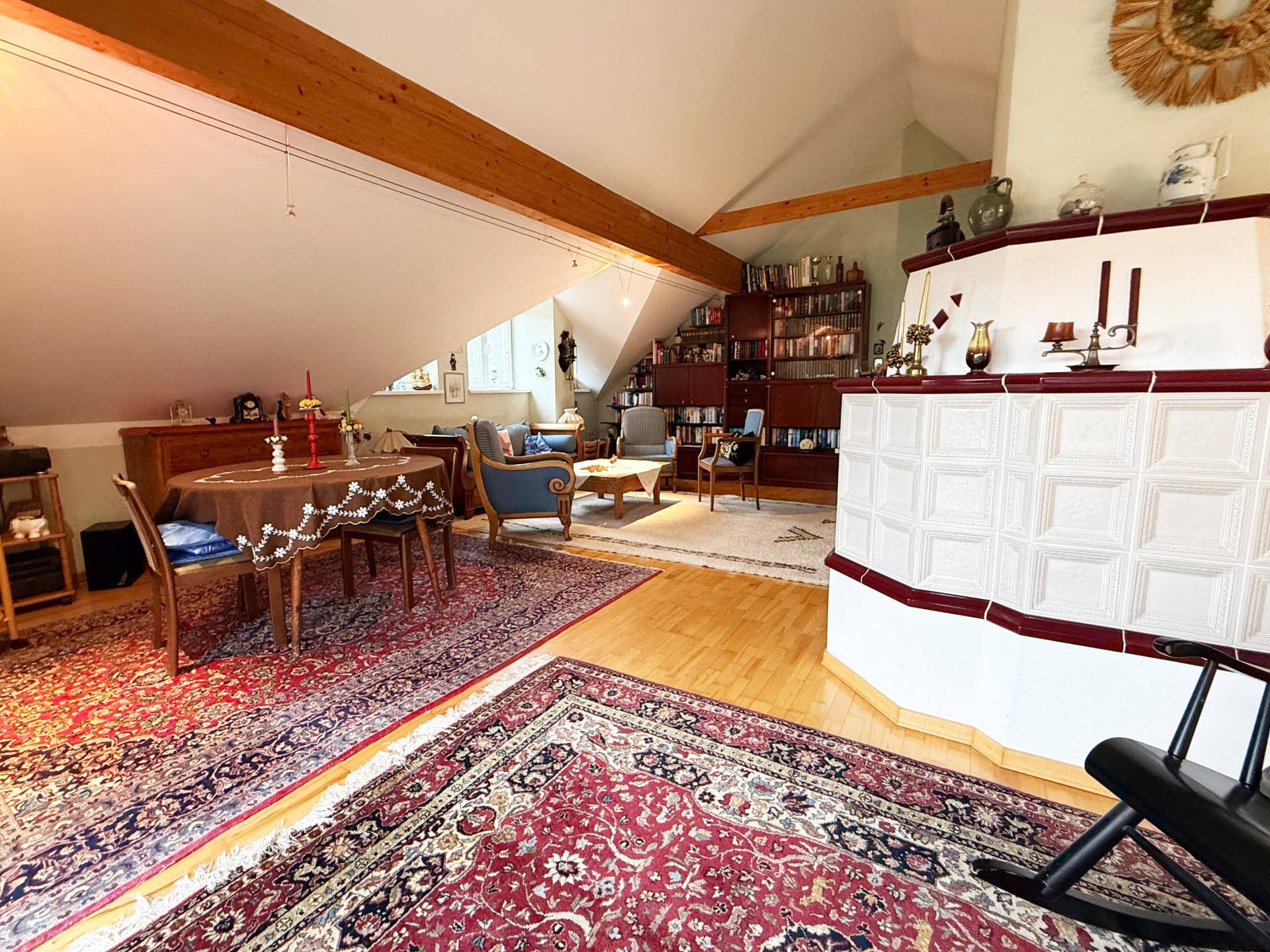 Geräumiges Wohnzimmer im Dachgeschoss mit Holzbalken, Kachelofen und traditionellen Teppichen.