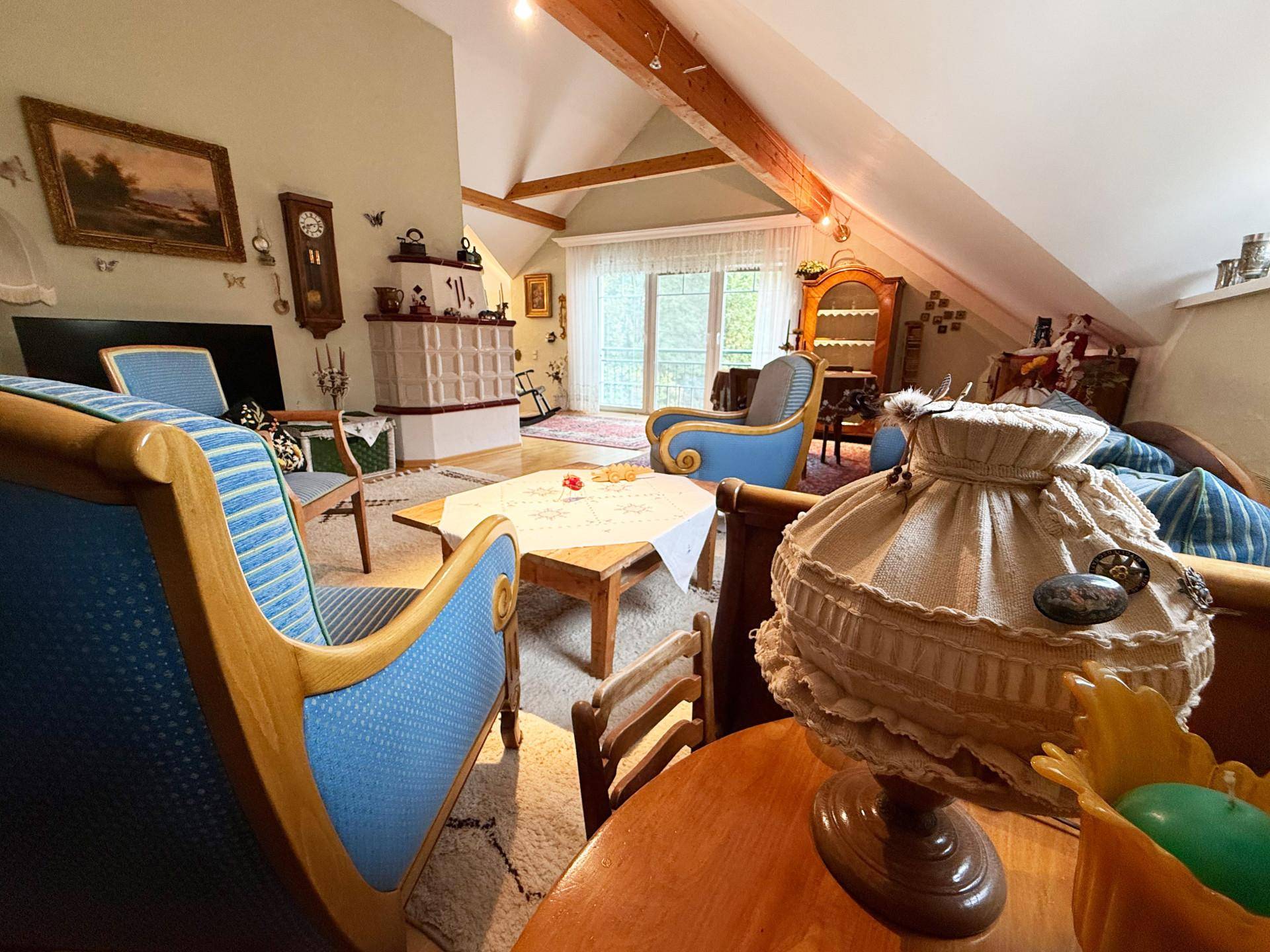 Helles Wohnzimmer mit traditionellem Kachelofen, Holzbalken und Zugang zu einem Außenbereich.