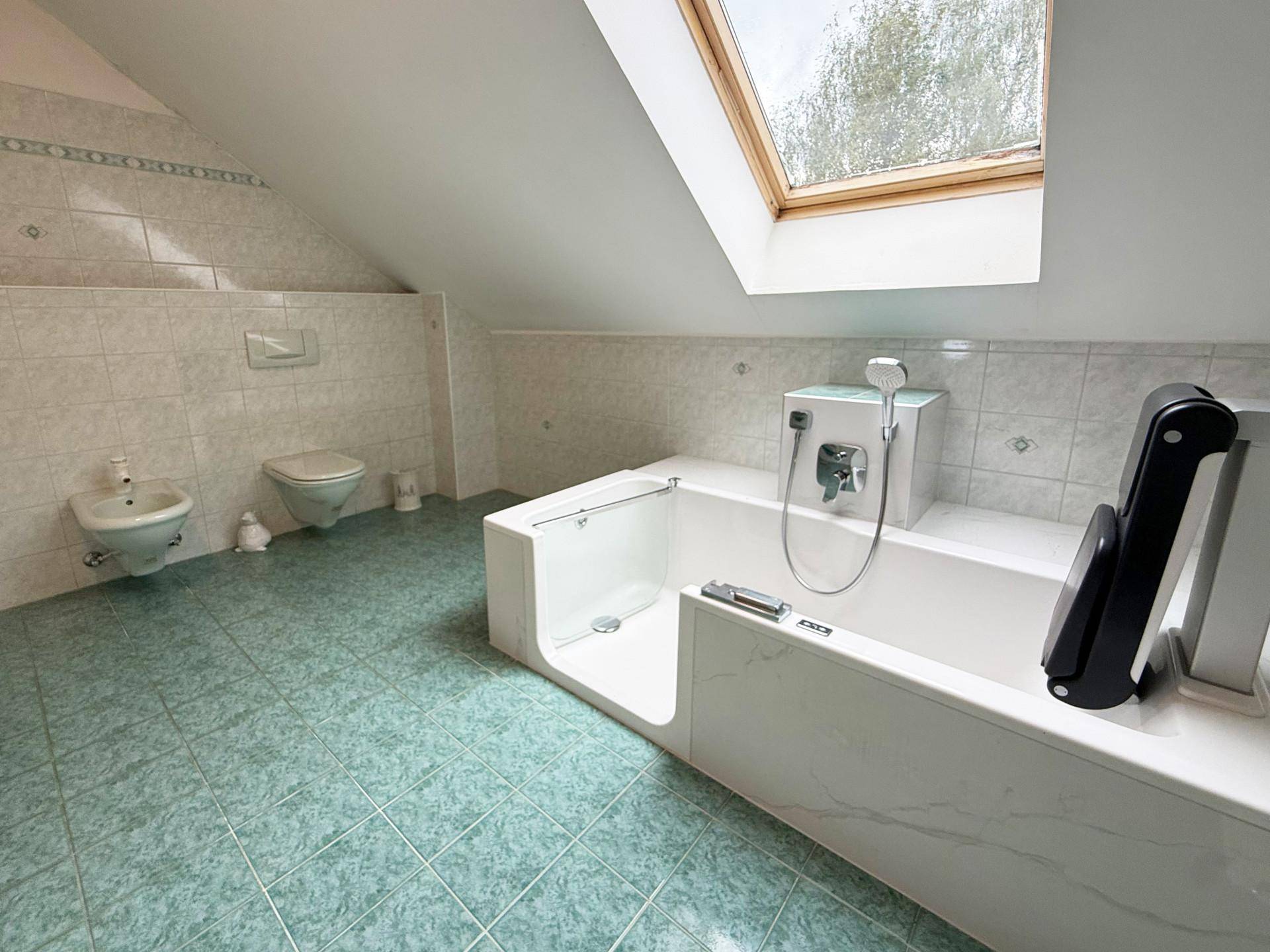 Helles Badezimmer mit Badewanne, Bidet, wandhängendem WC und grün gefliestem Boden unter einem Dachfenster.