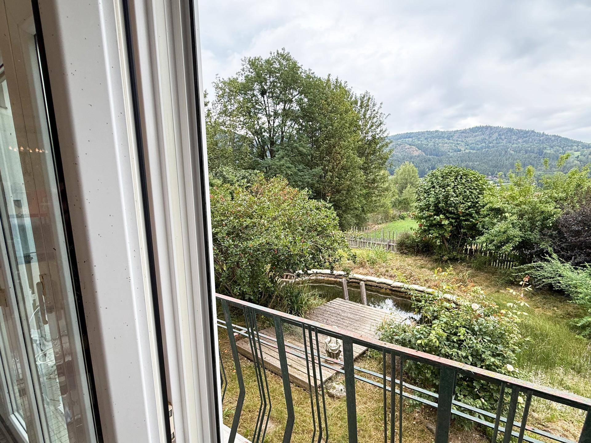 Blick aus dem Fenster auf einen naturnahen Garten mit Teich und grüner Berglandschaft.