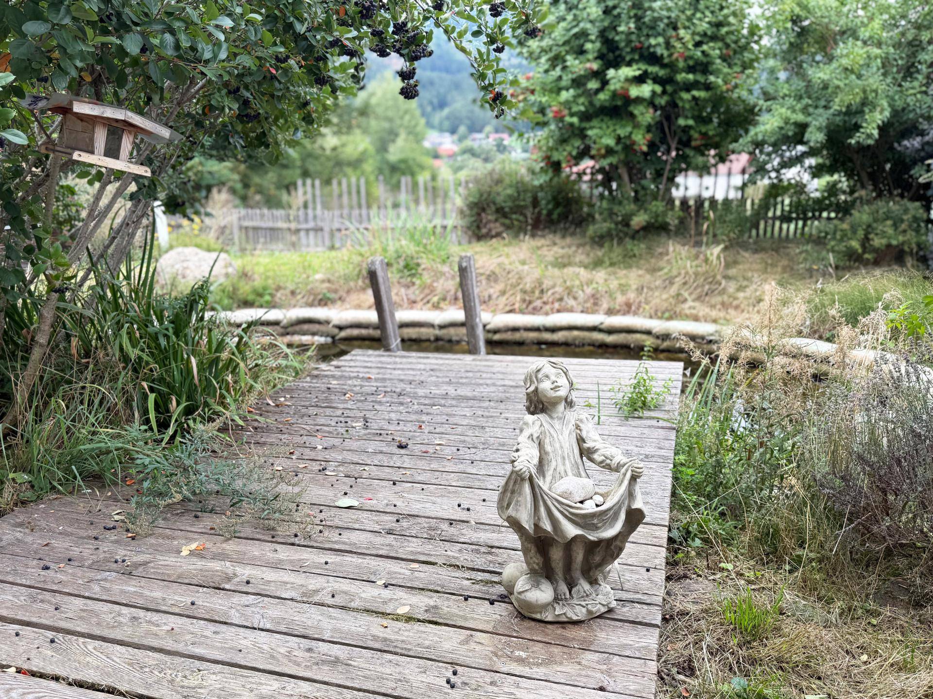 Holzsteg im Garten mit einer Kinderstatue, Vogelhäuschen und Blick auf den Teich.