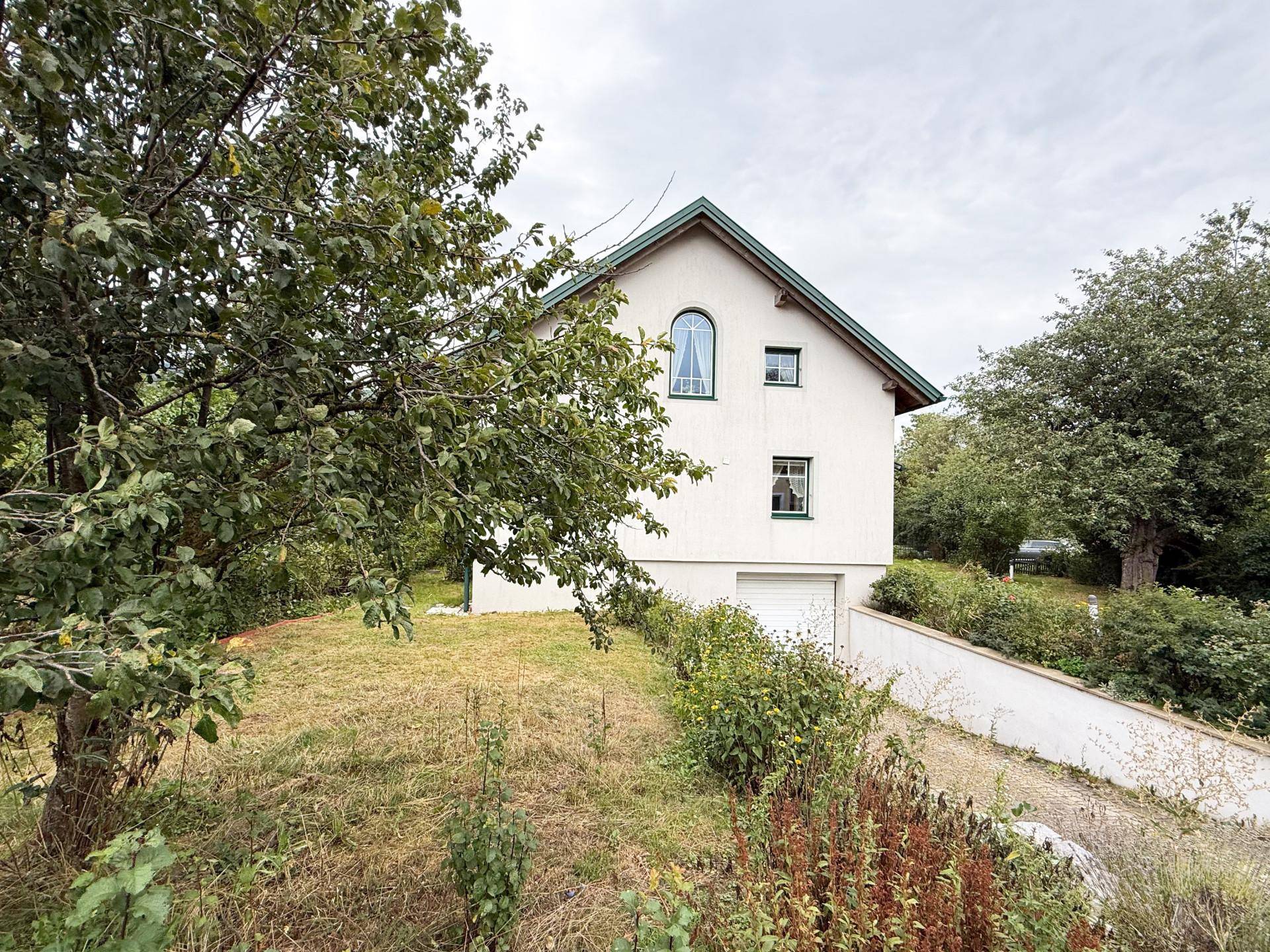 Weißes Einfamilienhaus mit grünem Satteldach, integrierter Garage und umgebender Natur.