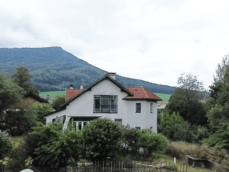 Freistehendes Haus mit rotem Ziegeldach, Balkon und umgeben von Bäumen vor einer Bergkulisse.
