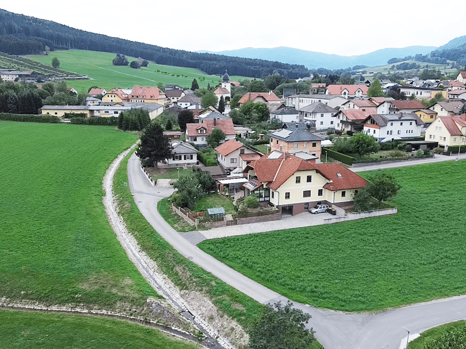 Weitläufige Luftaufnahme eines ländlichen Dorfes, umgeben von grünen Feldern und Bergen.