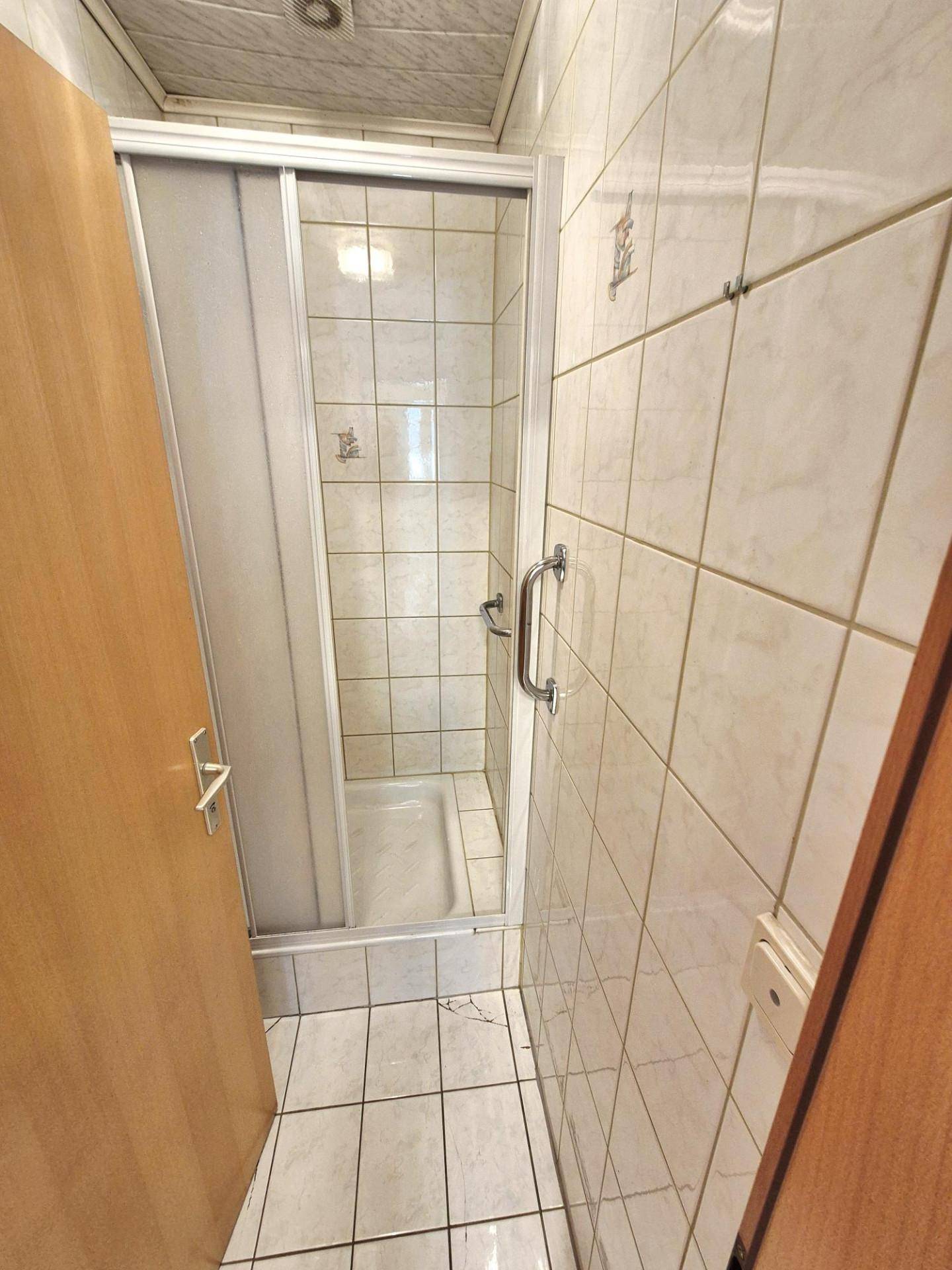 Kompaktes Badezimmer mit Duschkabine, weißen Fliesen und Haltegriff für mehr Sicherheit.