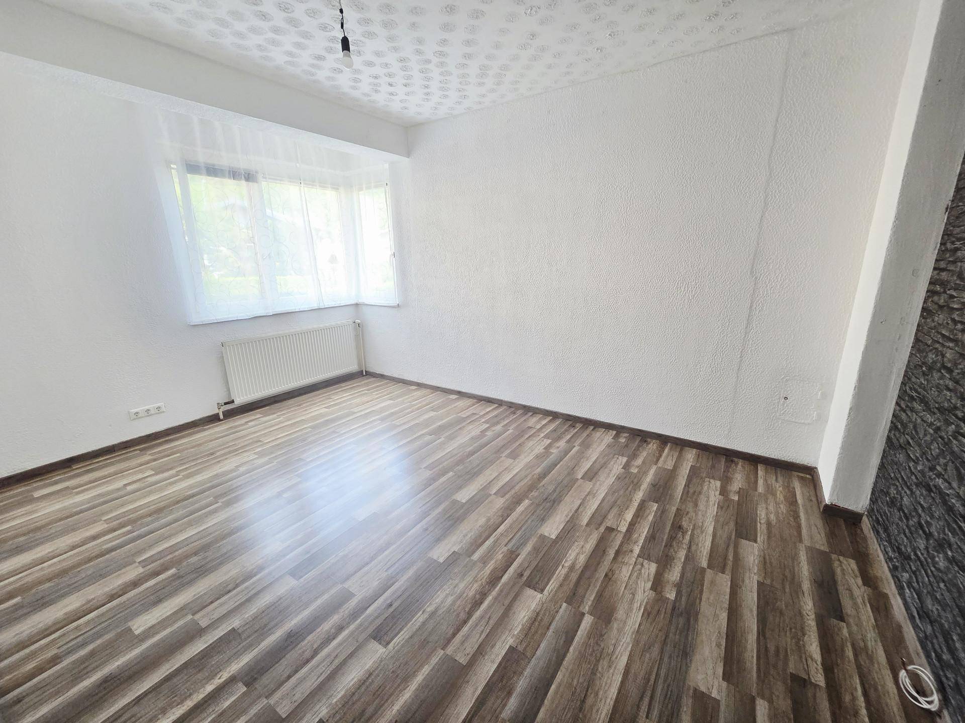 Helles, leeres Zimmer mit Laminatboden, weißen Wänden und einem Fenster mit Vorhängen.
