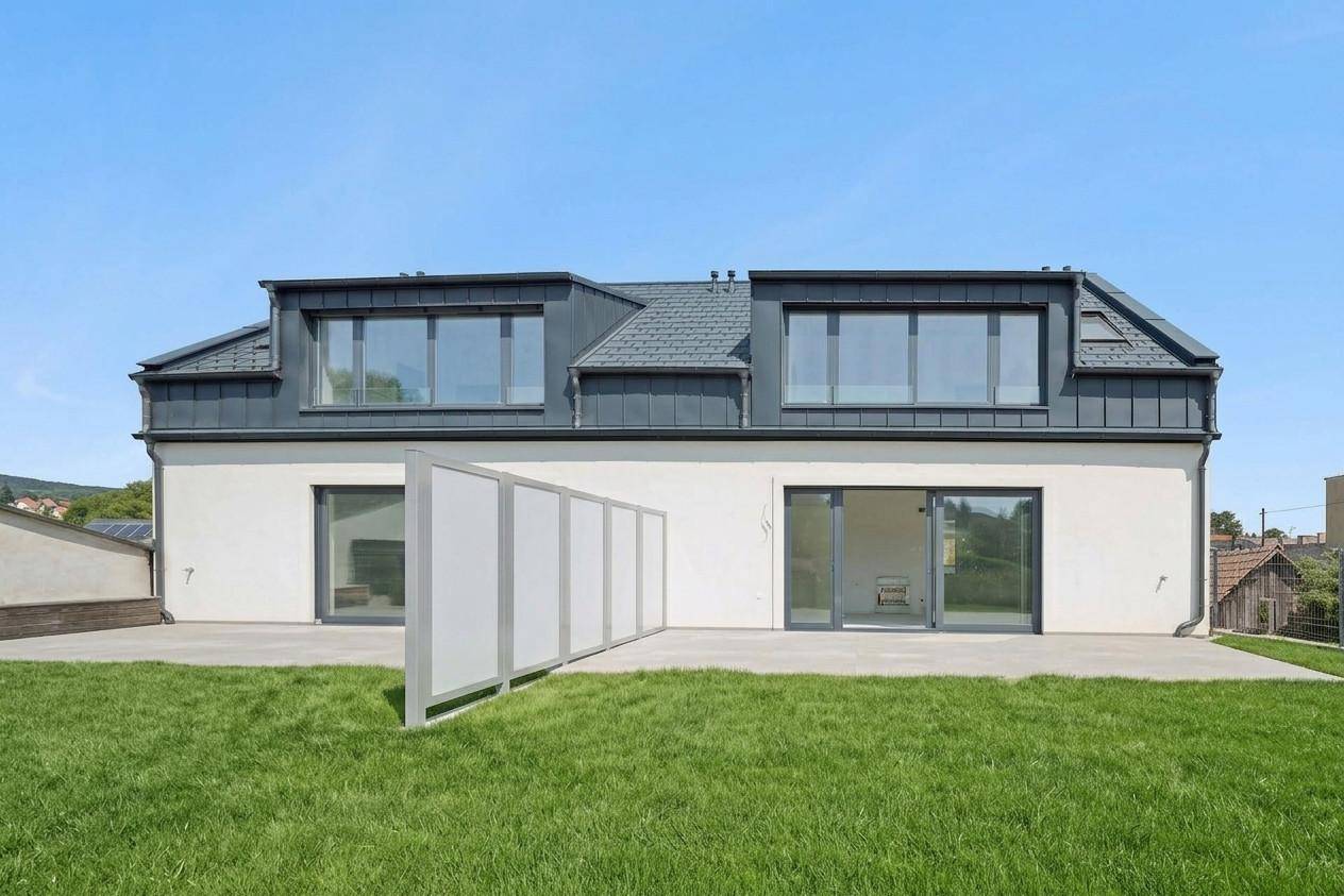 Rückseite eines modernen Gebäudes mit großer, unmöblierter Terrasse und angrenzender Rasenfläche.