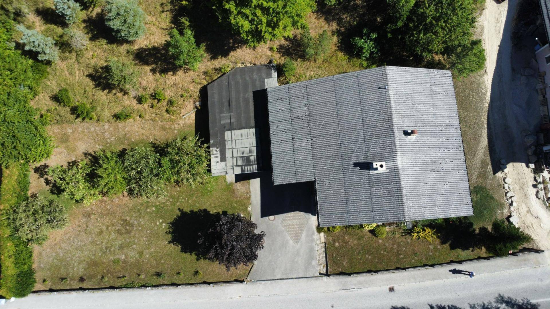 Luftaufnahme eines Hauses mit grauem Dach und angrenzendem Grundstück, umgeben von Bäumen.