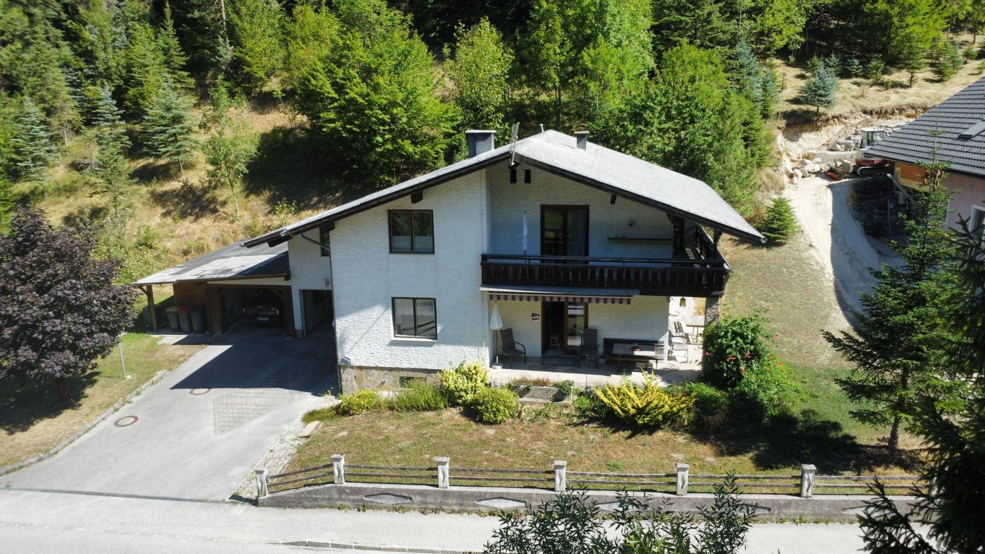 Traditionelles Haus mit Satteldach, Balkon und Carport, umgeben von viel Grün.