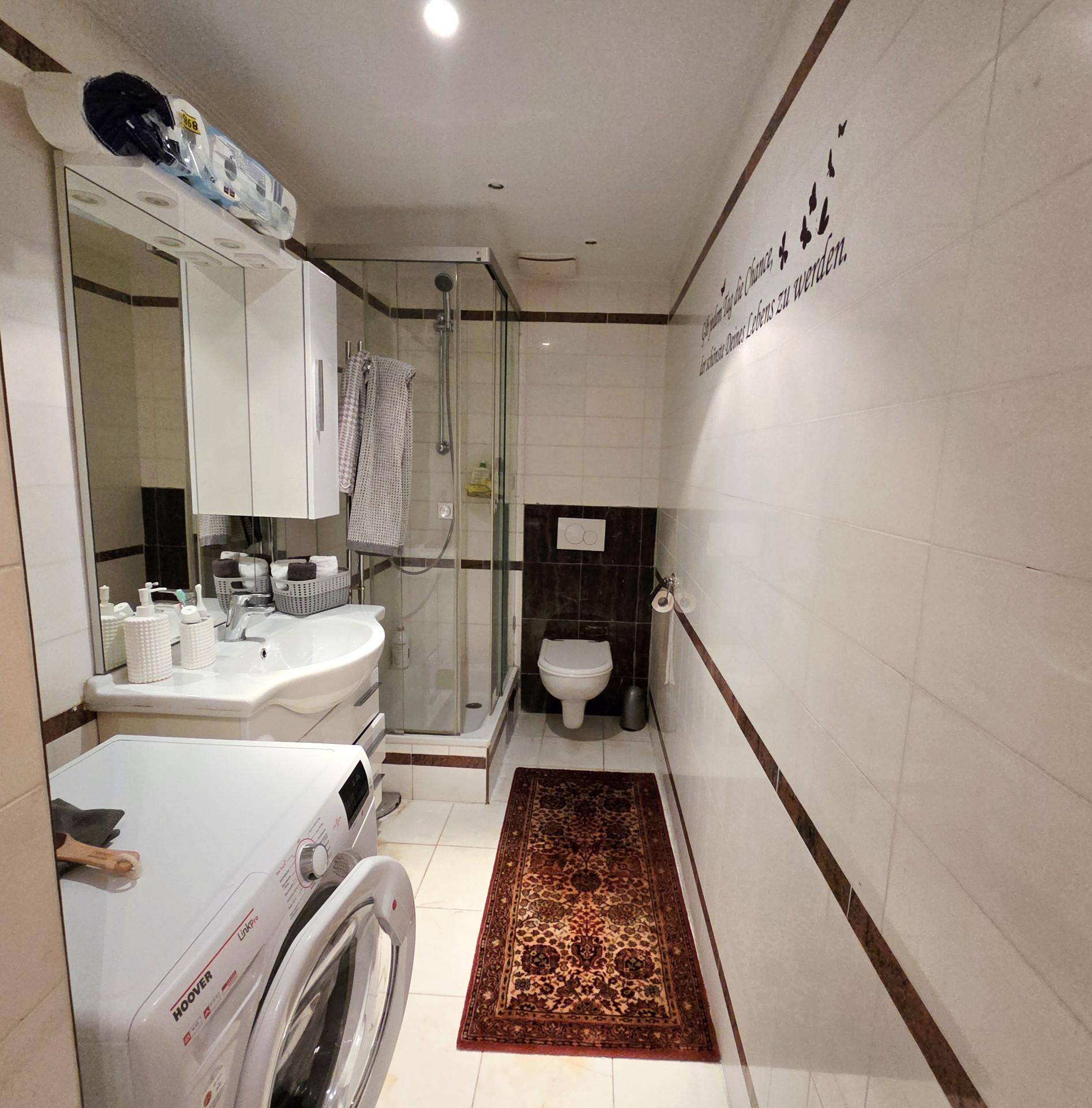 Schmaler Badezimmer mit Dusche, WC, Waschbecken und Waschmaschine, ausgestattet mit weißen Fliesen.
