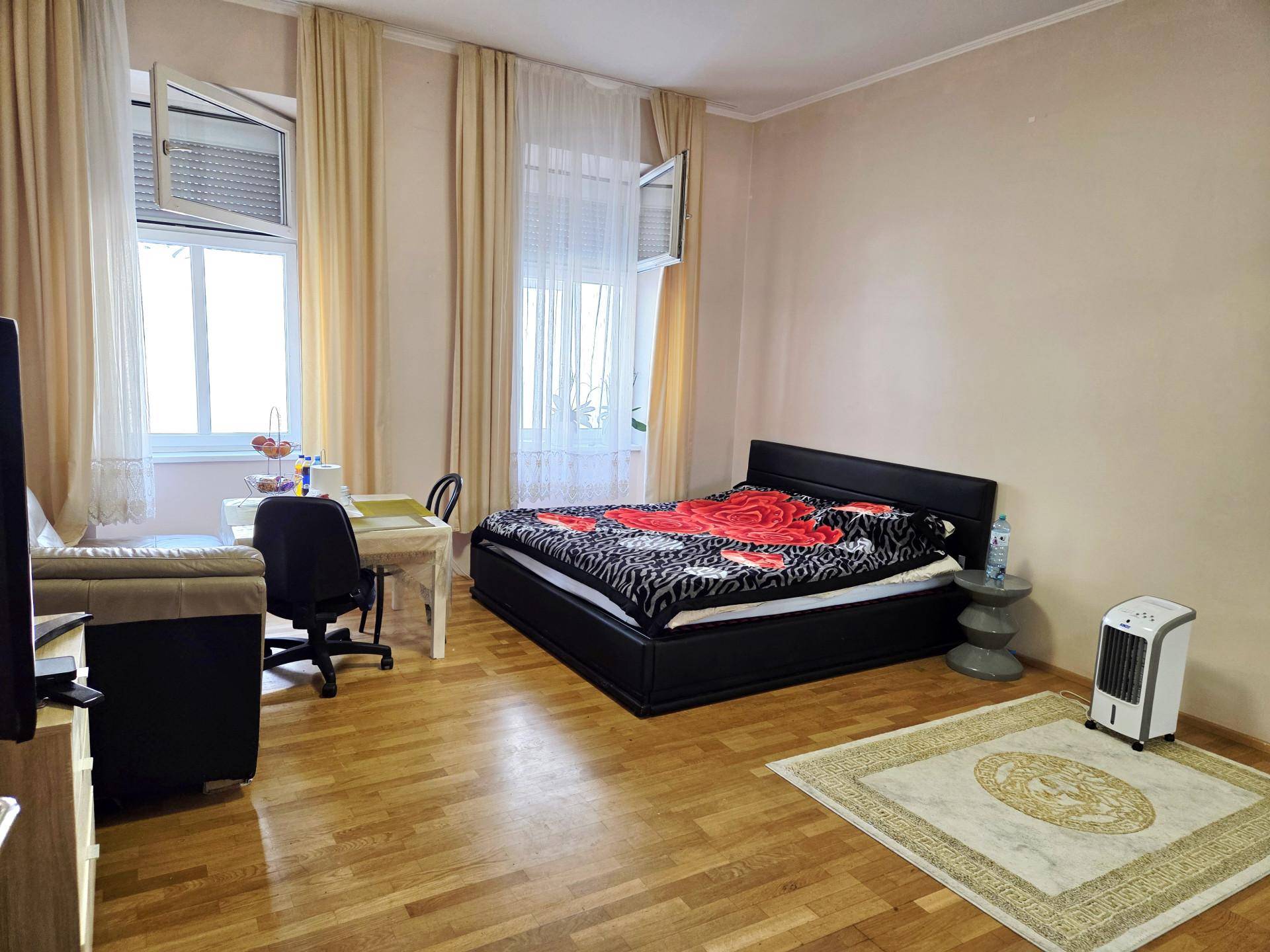 Kombinierter Wohn- und Schlafbereich mit Holzboden, einem Bett, Sofa und Schreibtisch, hell durch große Fenster.