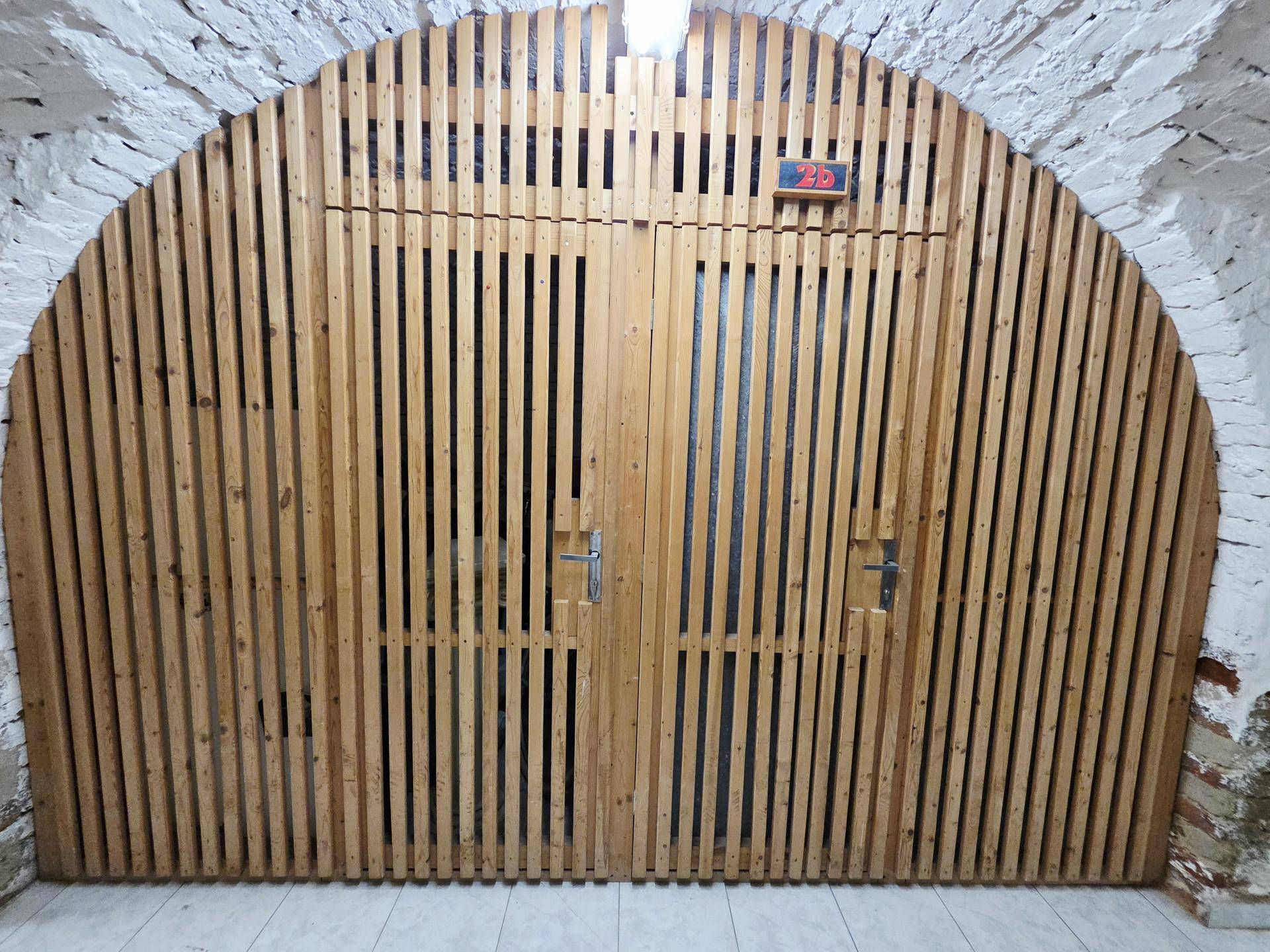 Gewölbte Holztüren zu Lagerabteilen in einem Kellerraum mit weiß getünchten Ziegelwänden.