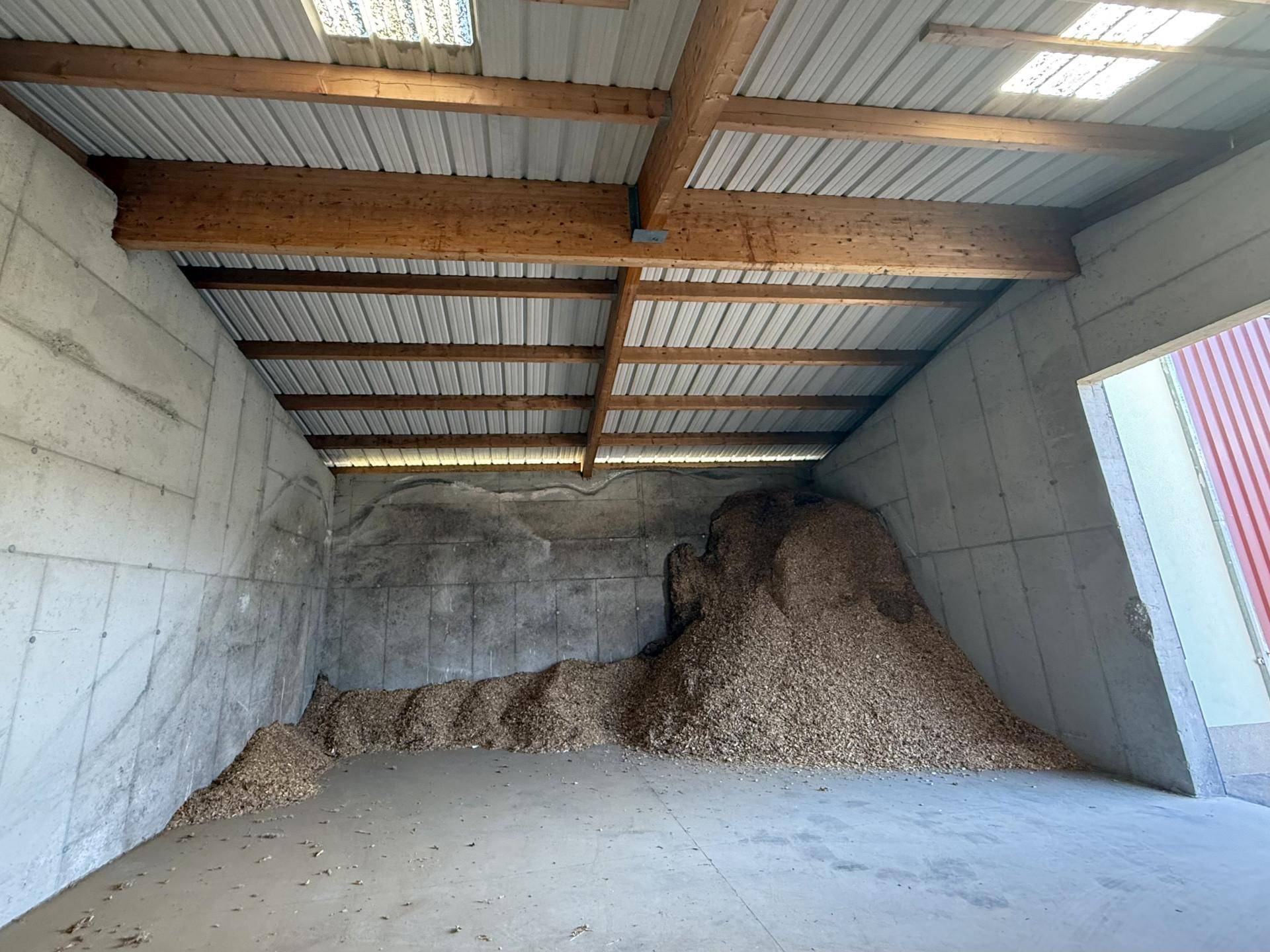 Große Lagerhalle mit Betonwänden, Holzbalken und einem Berg Hackschnitzel unter dem Wellblechdach.