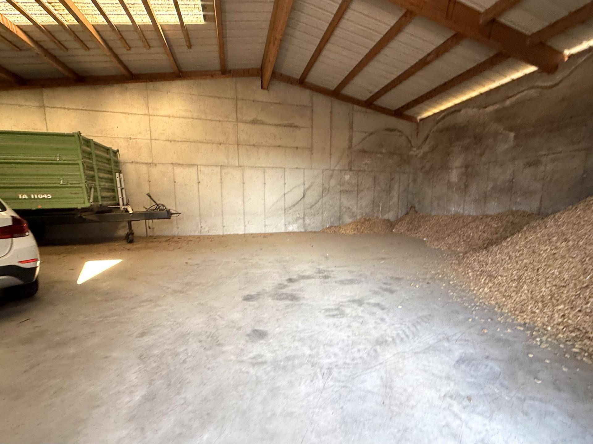 Innenansicht einer Lagerhalle mit Betonboden, Holzbalken und einem grünen Anhänger neben Hackschnitzeln.