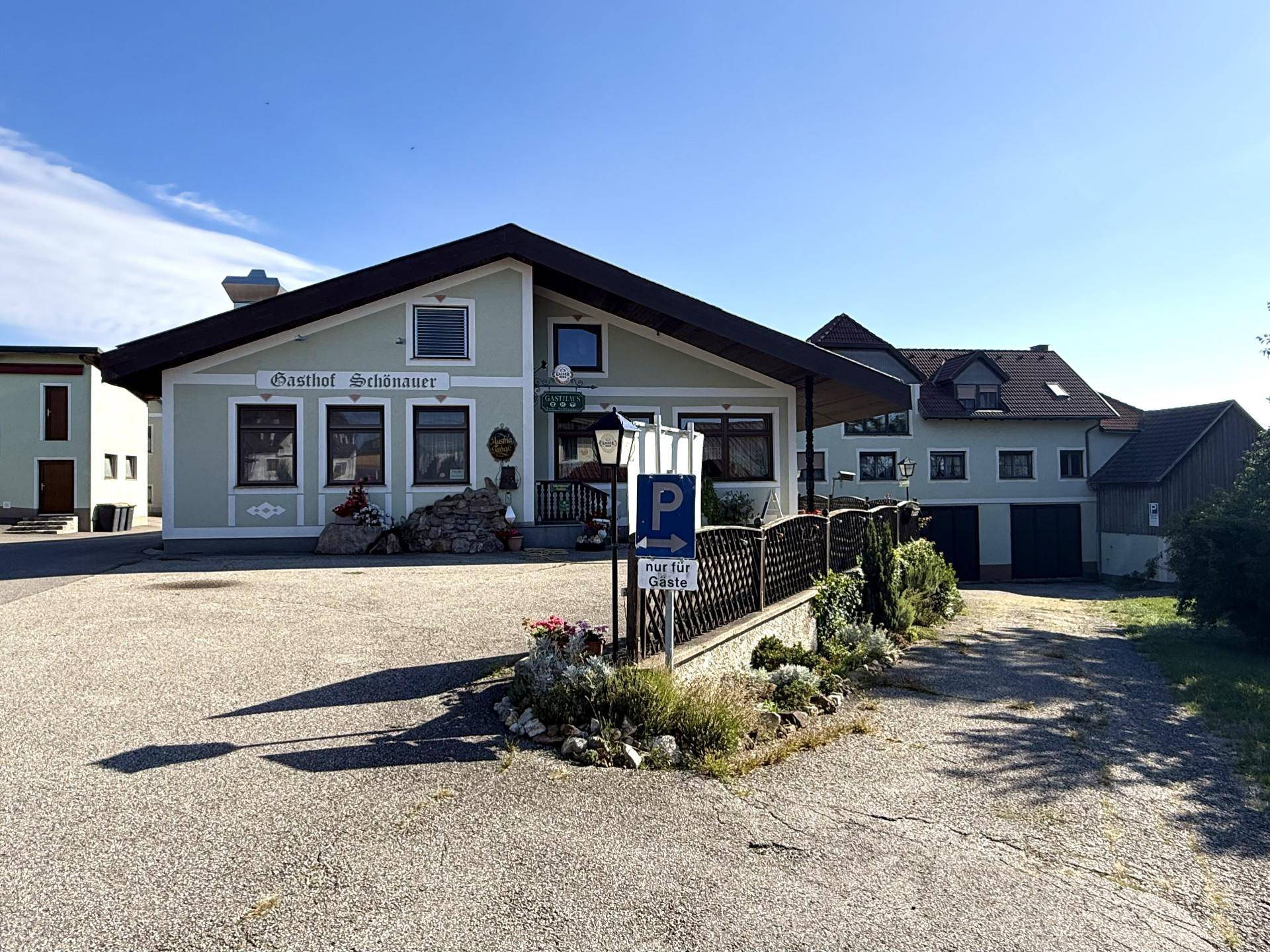 Vorderansicht des Gasthofs Schönauer mit hellgrüner Fassade, dunklem Dach und einem Schotterparkplatz.
