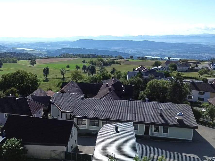 Panoramische Luftaufnahme des Dorfes und der umliegenden Landschaft mit fernen Bergen und einem See.