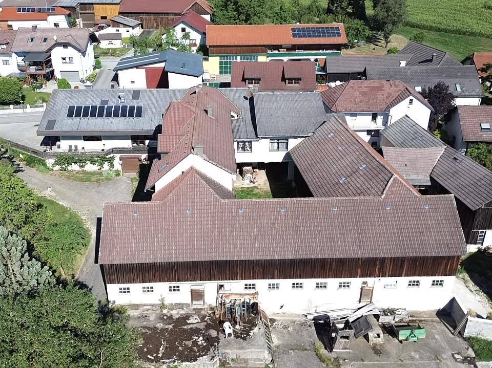 Luftaufnahme des langen Scheunengebäudes und weiterer Häuser, umgeben von grünen Feldern und ländlicher Bebauung.