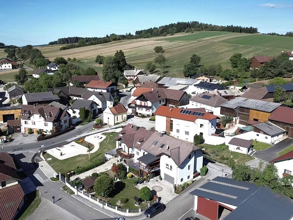 Weitläufige Luftaufnahme des Dorfes mit vielen Häusern, einer kleinen Kirche und sanften Hügeln im Hintergrund.