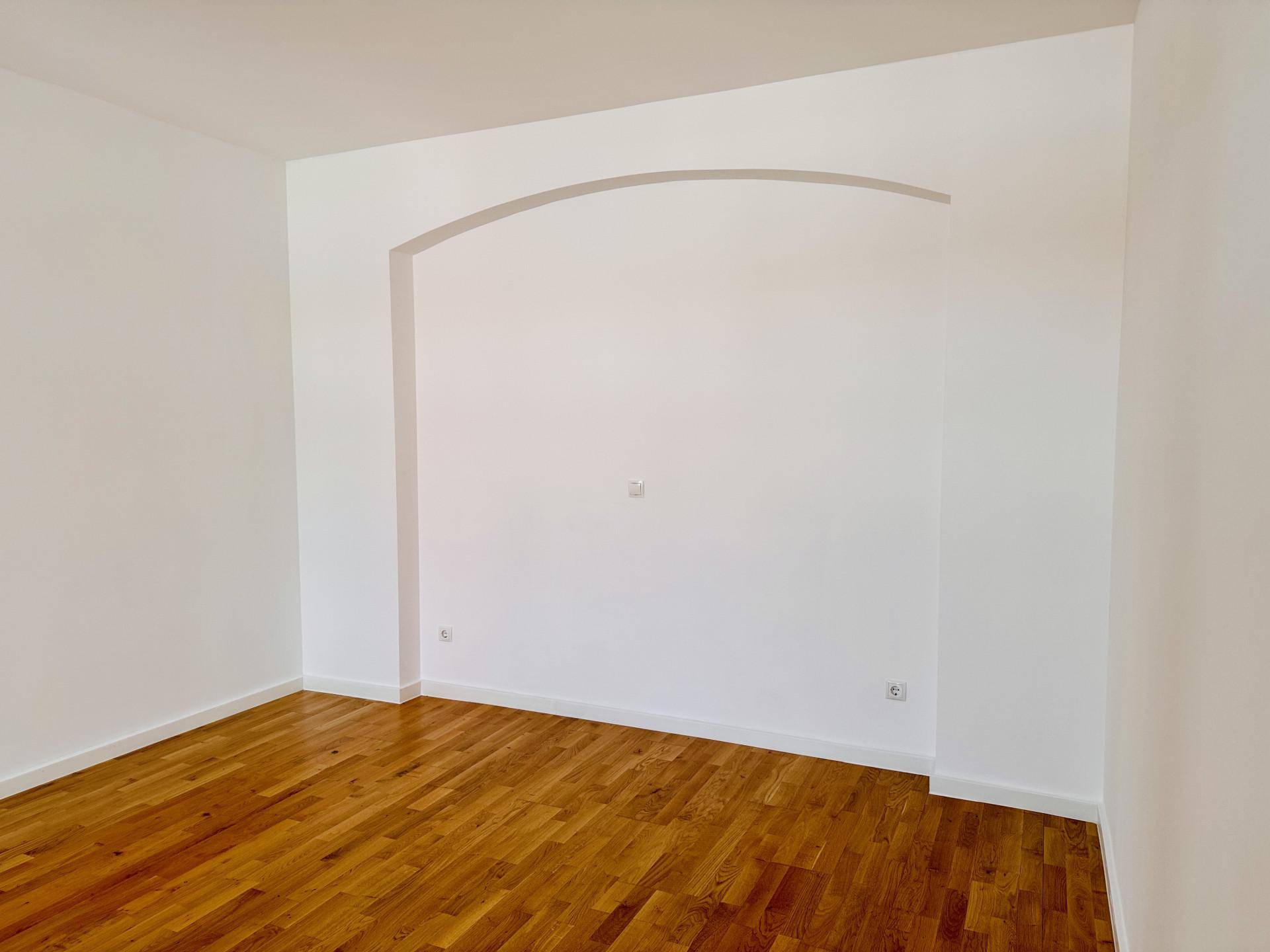 Leerer Raum mit warmem Holzboden und einer dekorativen Bogennische in der Wand.