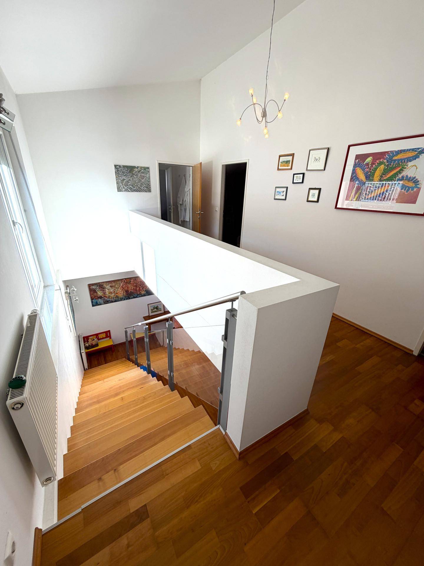 Heller Treppenaufgang mit Holzstufen, Glasgeländer und hohen Decken.