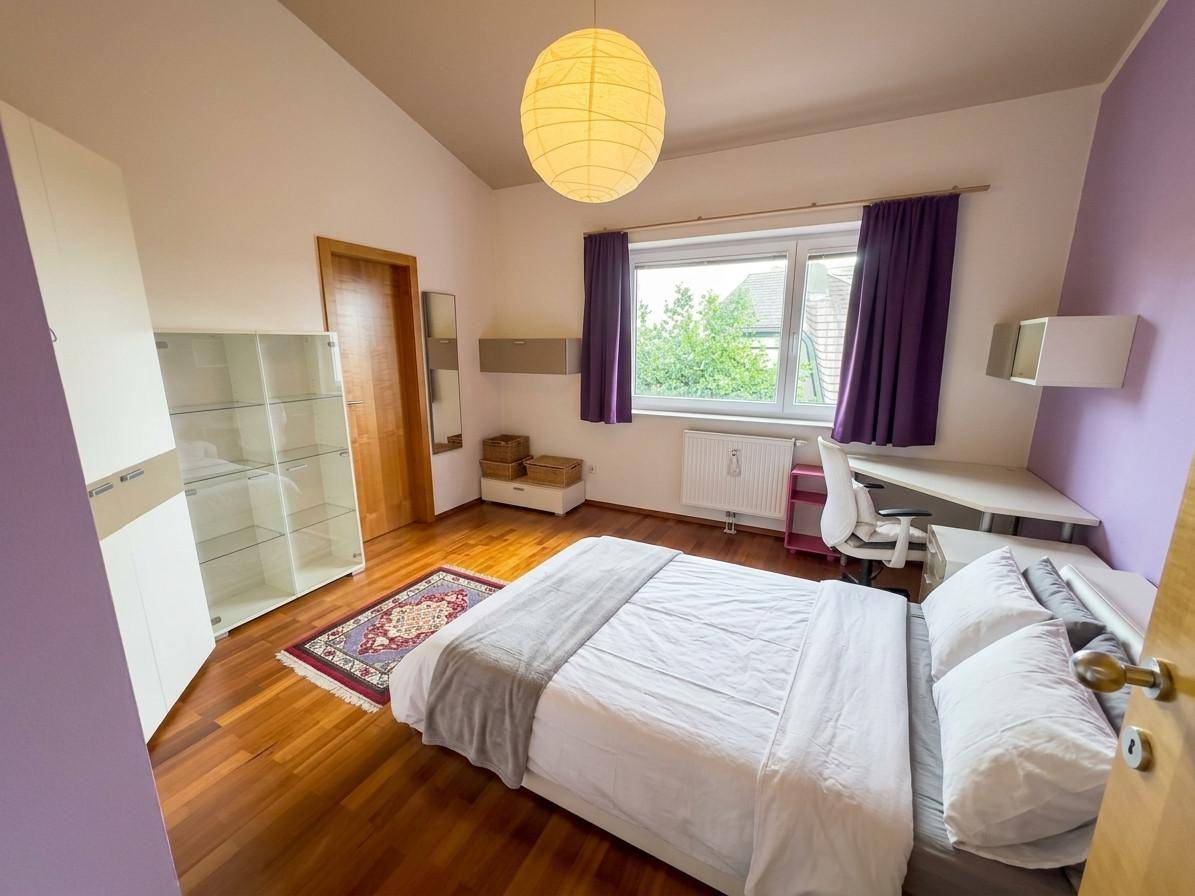 Schlafzimmer mit Dachschräge, Holzboden, Doppelbett und lila Vorhängen, bietet gemütlichen Wohnraum.