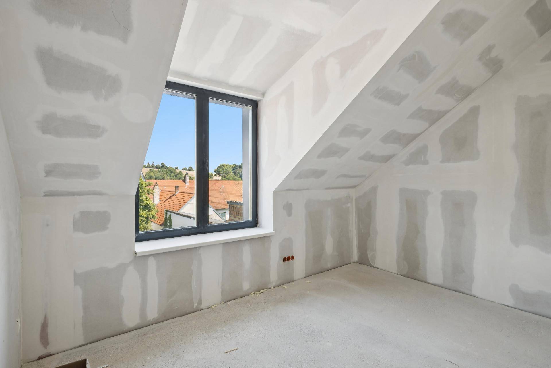 Dachgeschosszimmer im Rohbau mit schrägen Wänden und einem Fenster mit Blick auf Dächer.