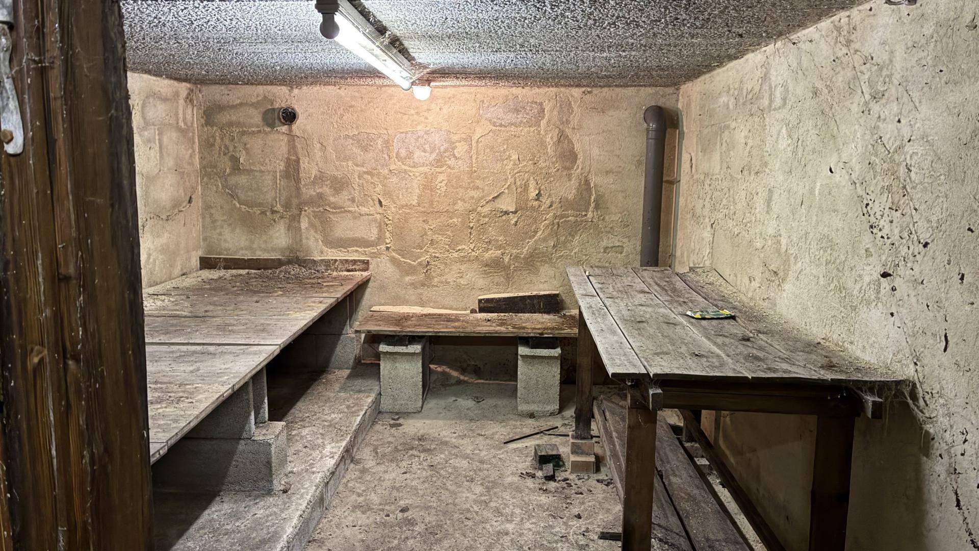 Rustikaler Kellerraum mit Holztischen und einfacher Beleuchtung, ideal als Werkstatt oder Lagerbereich.