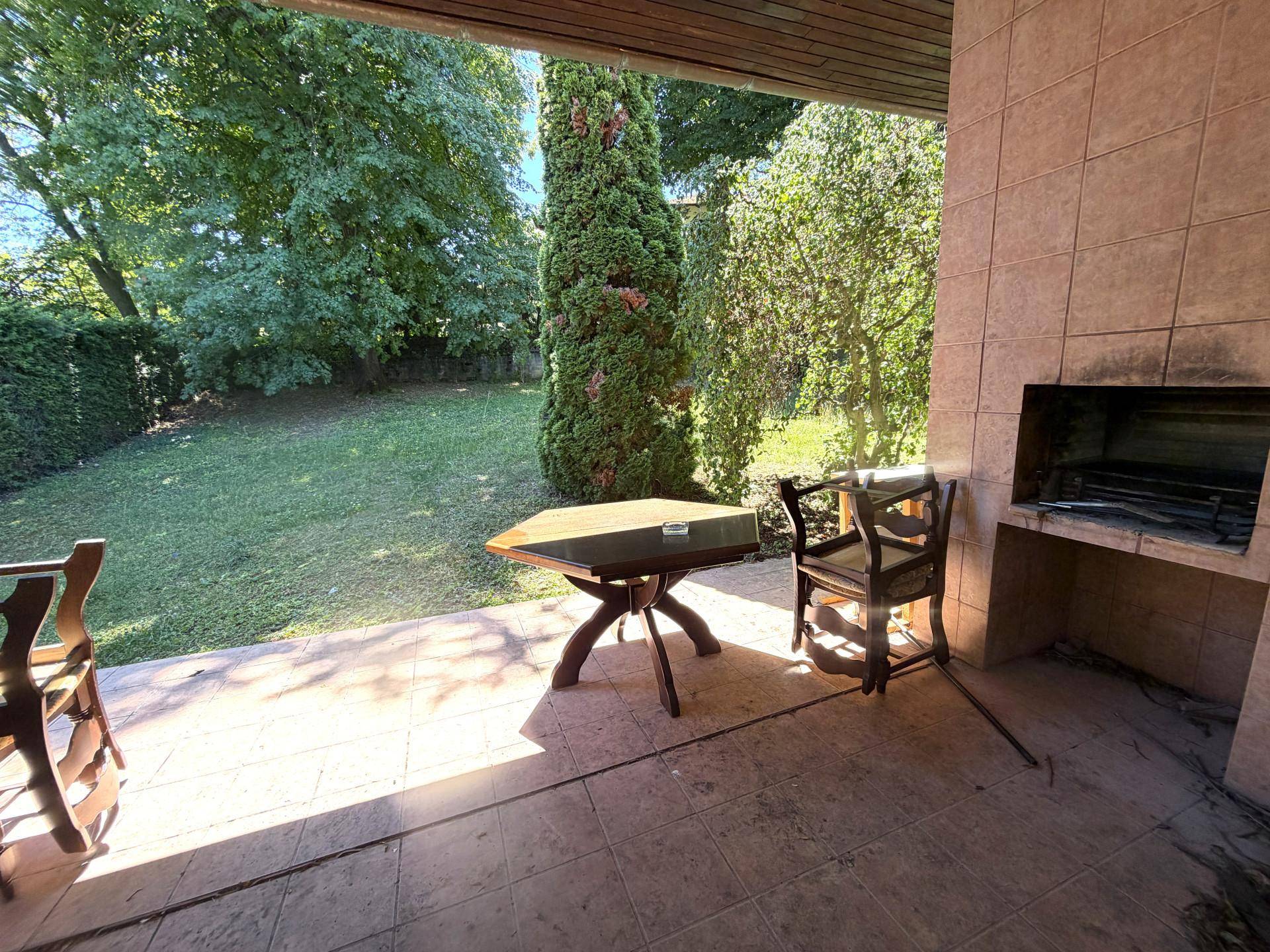 Überdachte Terrasse mit Außenkamin und Sitzgelegenheiten, umgeben von einem grünen Garten.