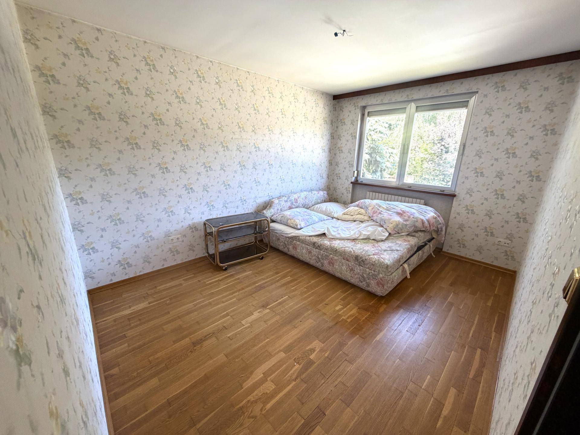 Schlafzimmer mit Holzboden, floraler Tapete und einem großen Fenster mit Blick ins Grüne.