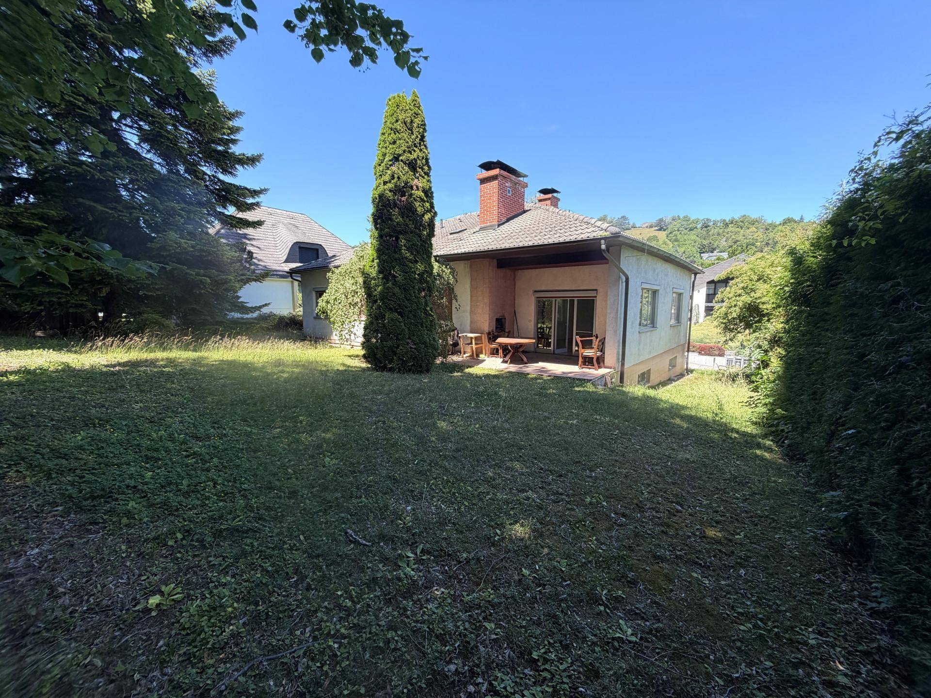 Rückseite des Hauses mit Terrasse und großem, sonnigen Gartenbereich, umgeben von Bäumen.