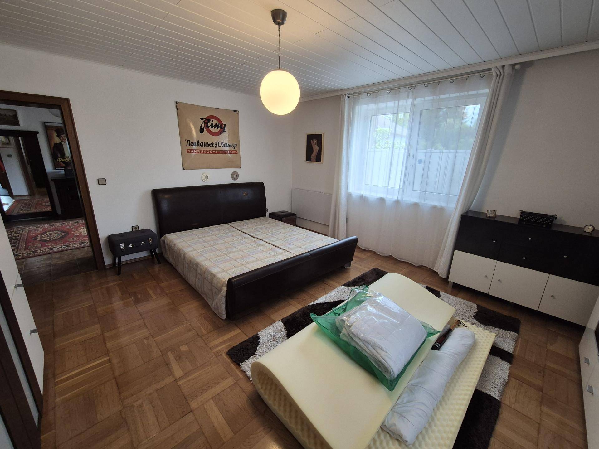 Schlafzimmer mit Parkettboden, Doppelbett und Fenster mit Vorhängen.