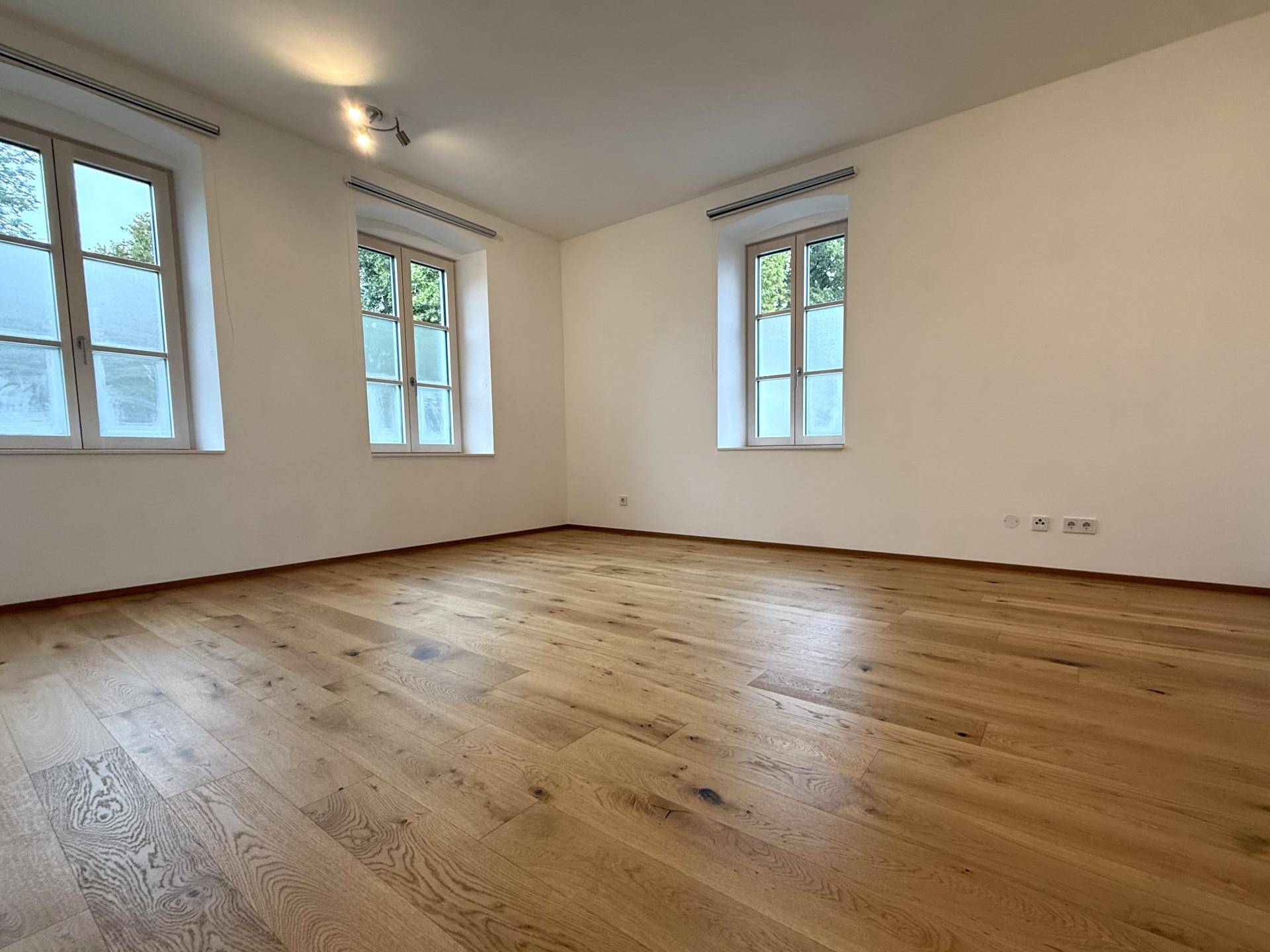 Heller, leerer Raum mit Holzboden und drei Fenstern, ideal als Wohn- oder Schlafzimmer.