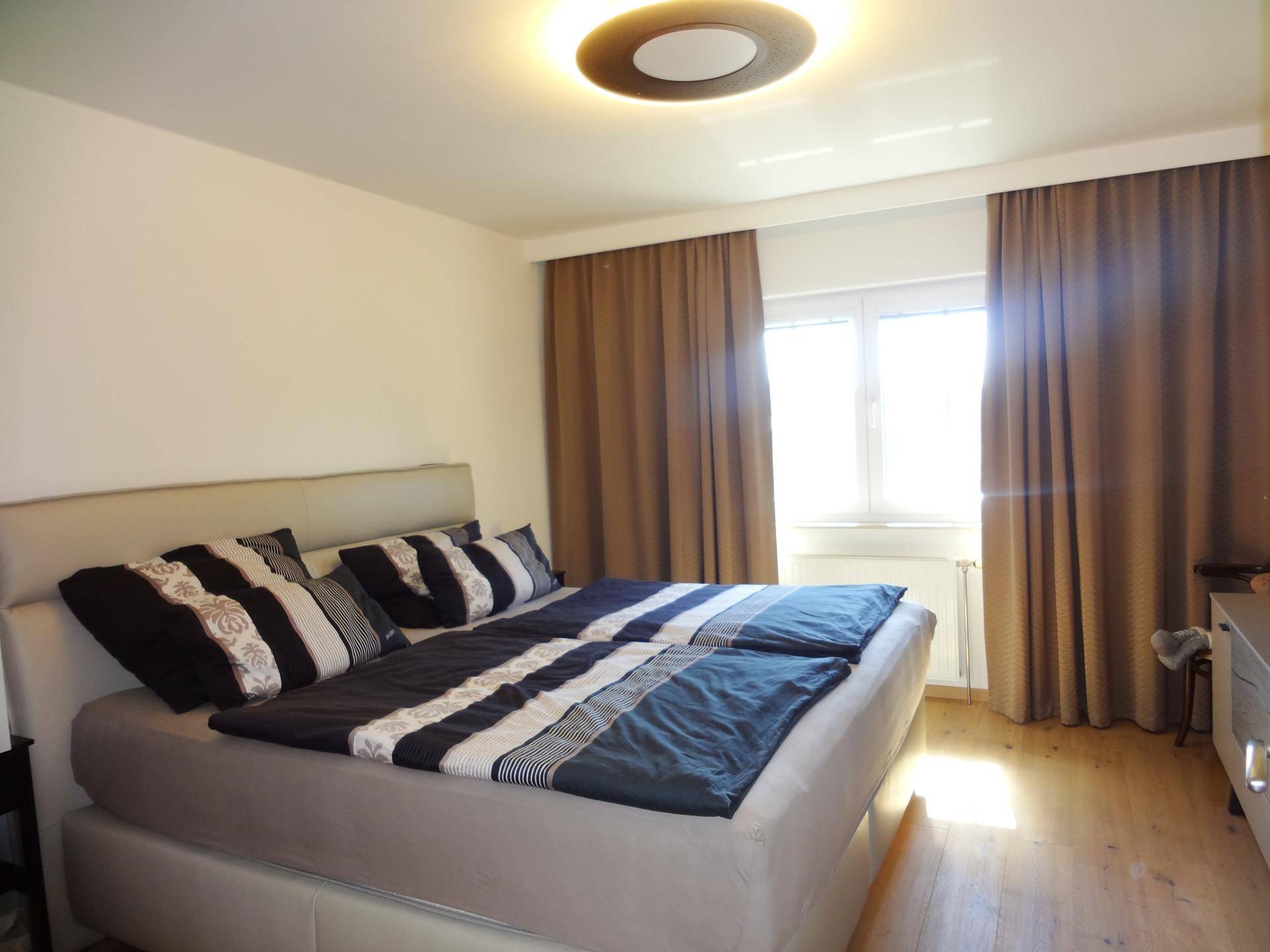 Helles Schlafzimmer mit Doppelbett, Holzboden und großen Fenstern mit braunen Vorhängen.