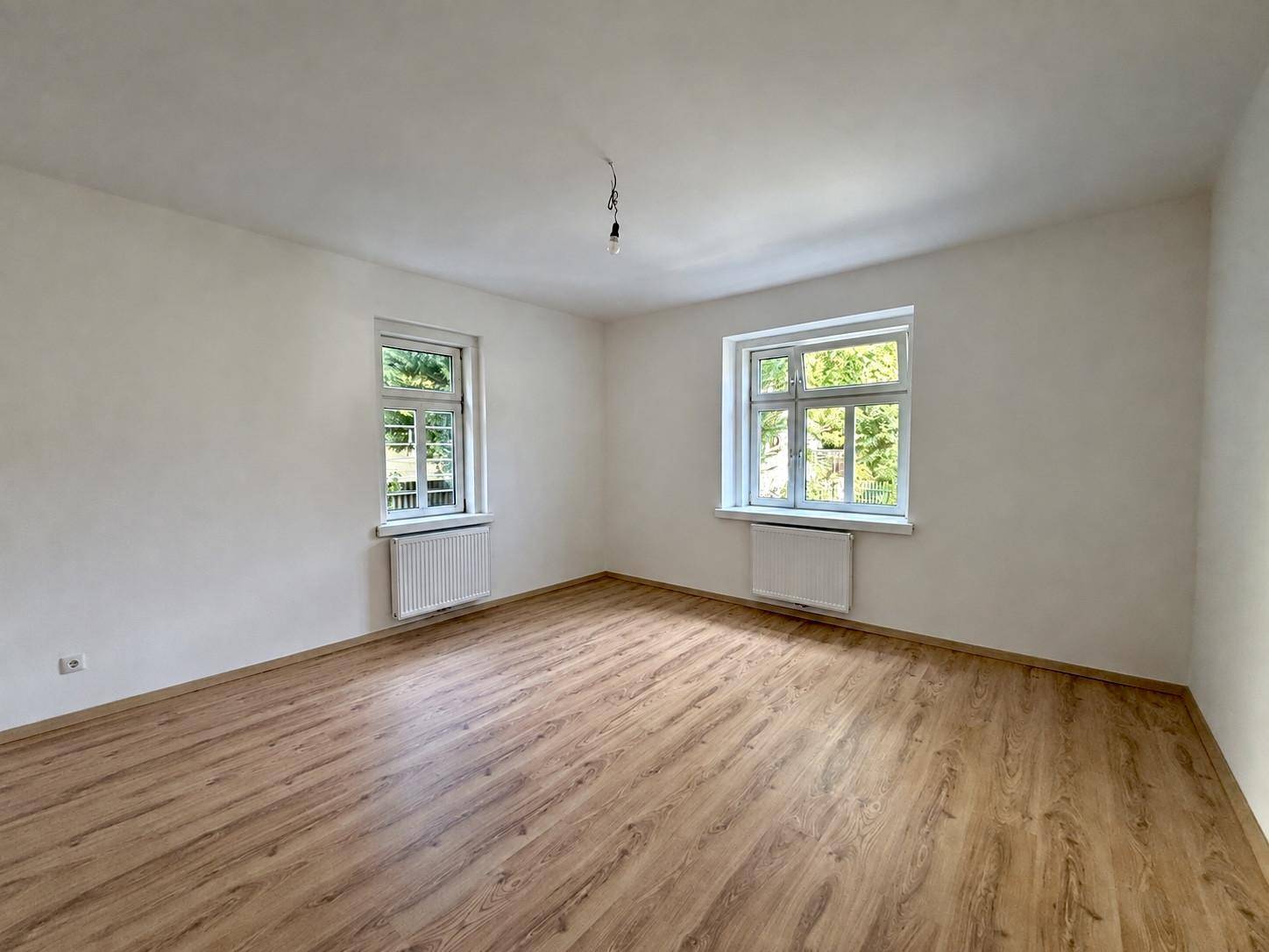 Helles, leeres Zimmer mit Laminatboden und zwei Fenstern, bereit zur individuellen Gestaltung.