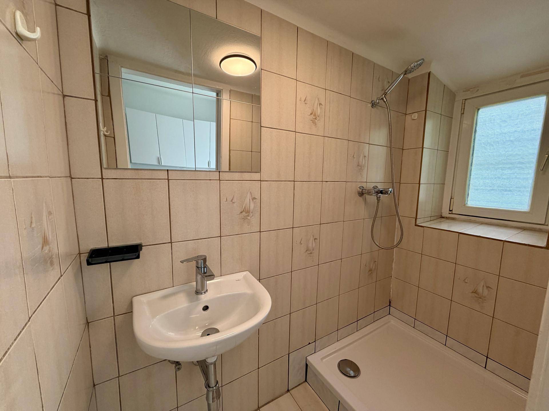 Gefliestes Badezimmer mit Dusche, Waschbecken und einem Fenster für Belüftung und Tageslicht.