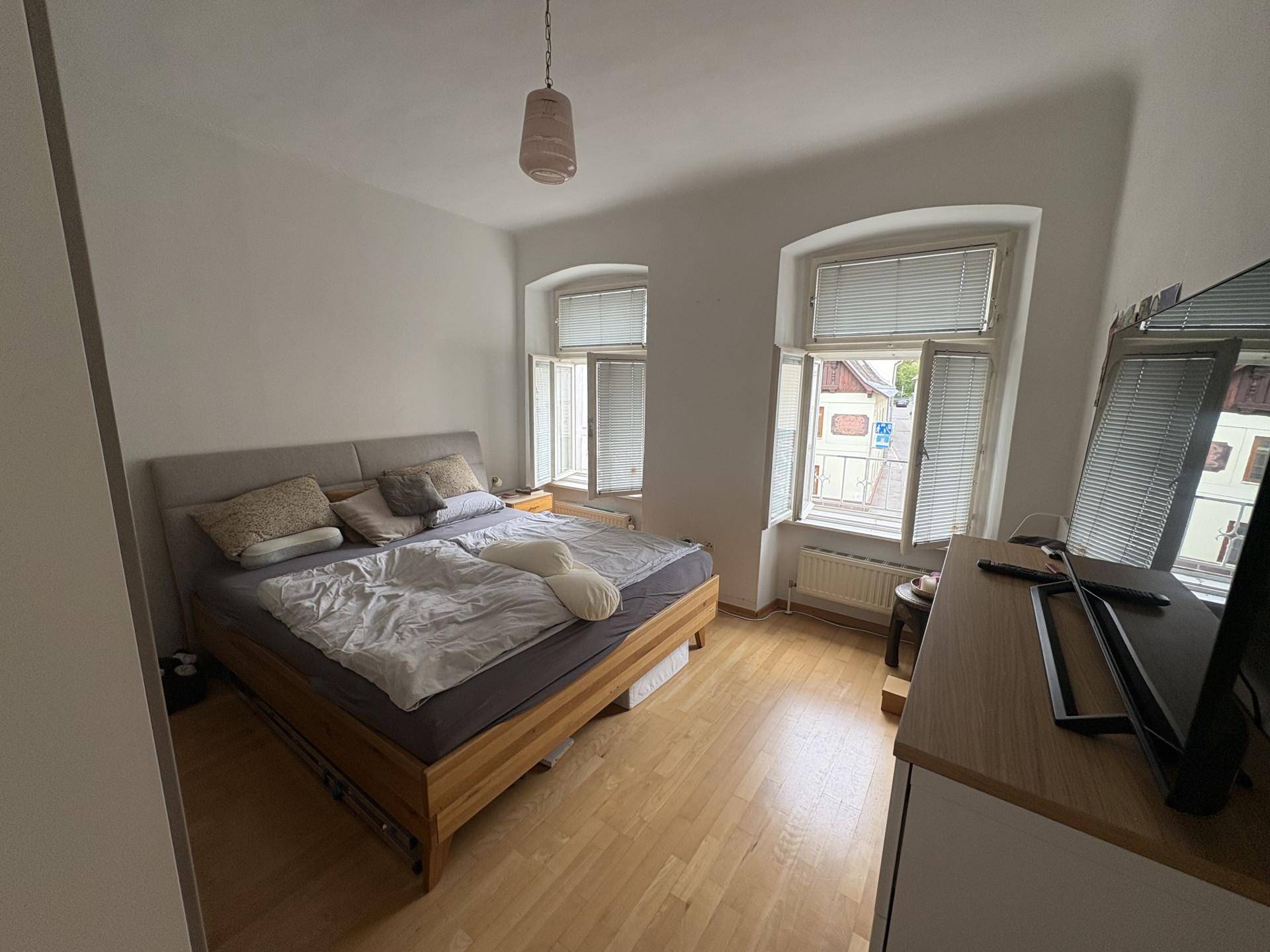 Gemütliches Schlafzimmer mit Doppelbett, Holzfußboden und zwei Fenstern zur Straße.
